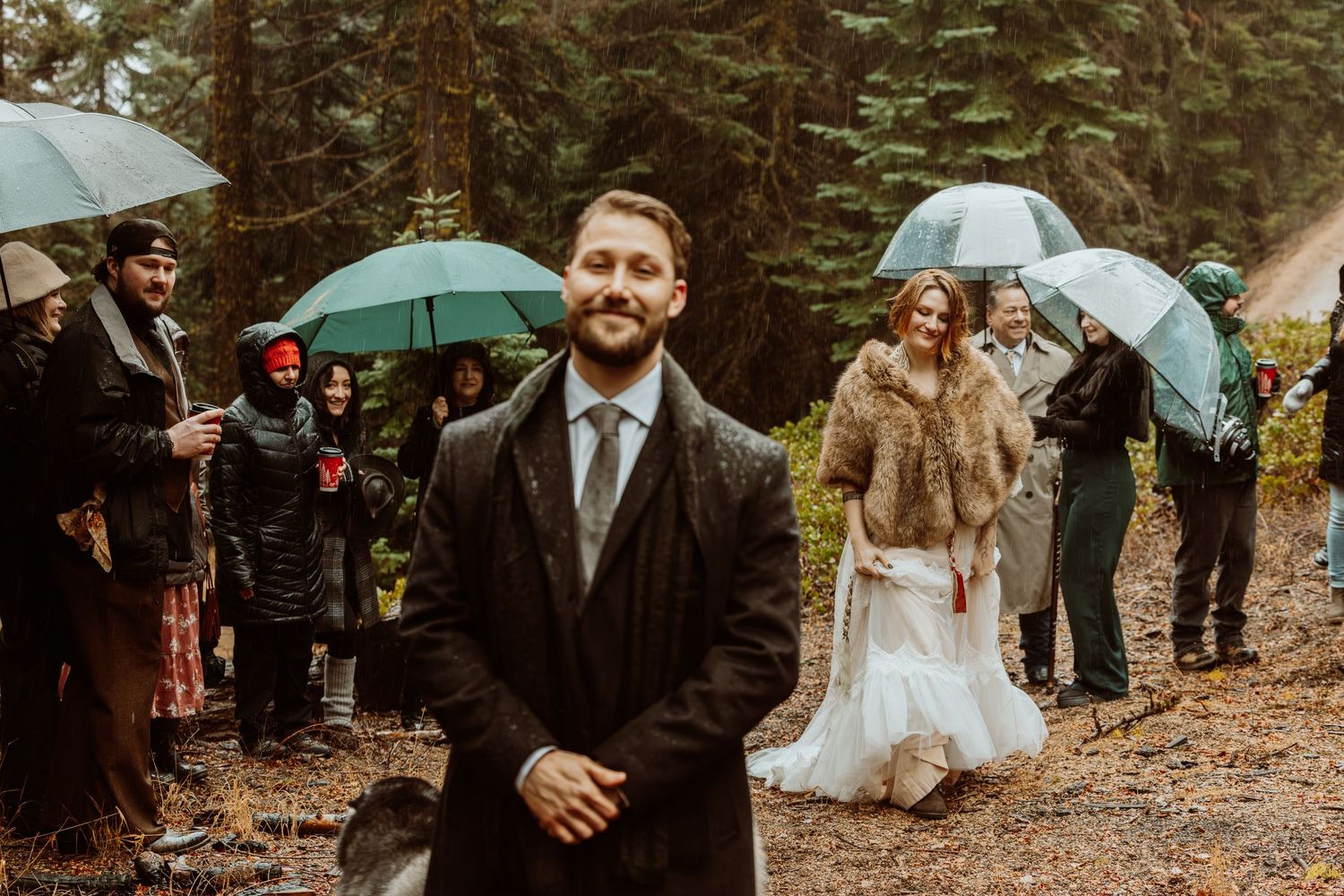 Groom in dark suit stands before bride in fur wrap with umbrella-holding guests in rainy forest.