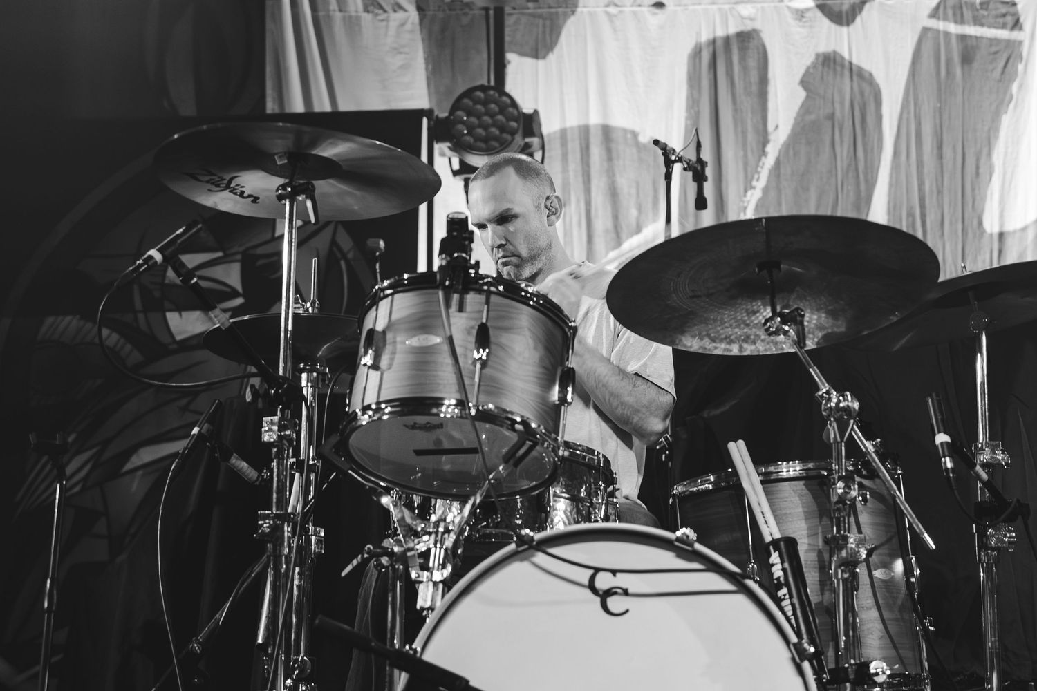 Black and white close-up of a focused drummer playing a full drum kit on a concert stage.