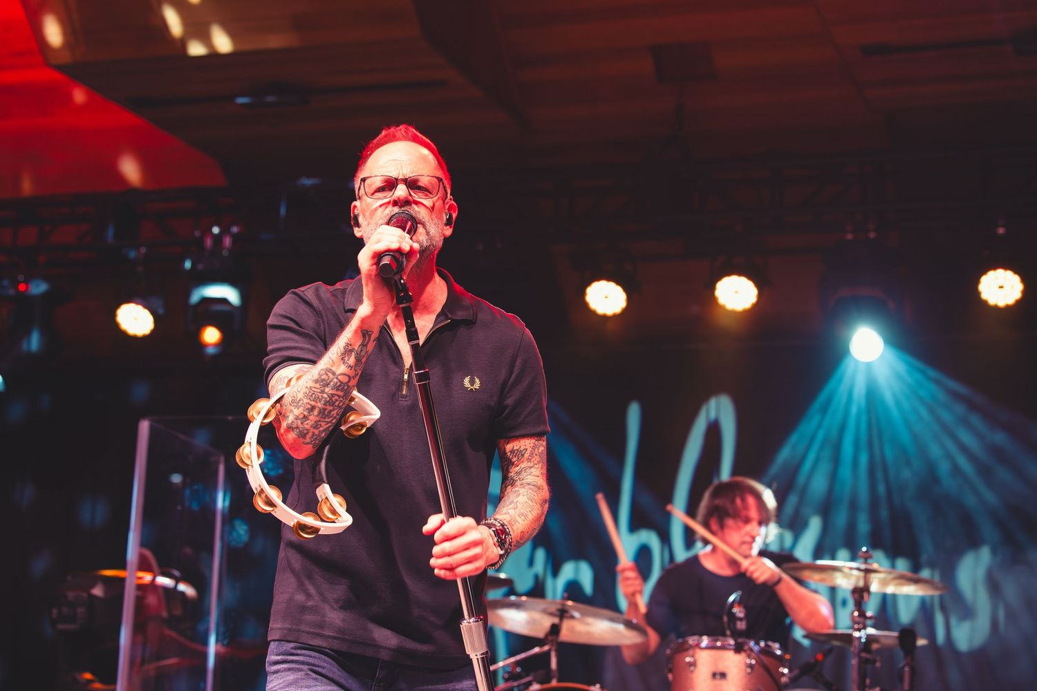 Tattooed lead singer performs holding a tambourine under blue and red concert lights.