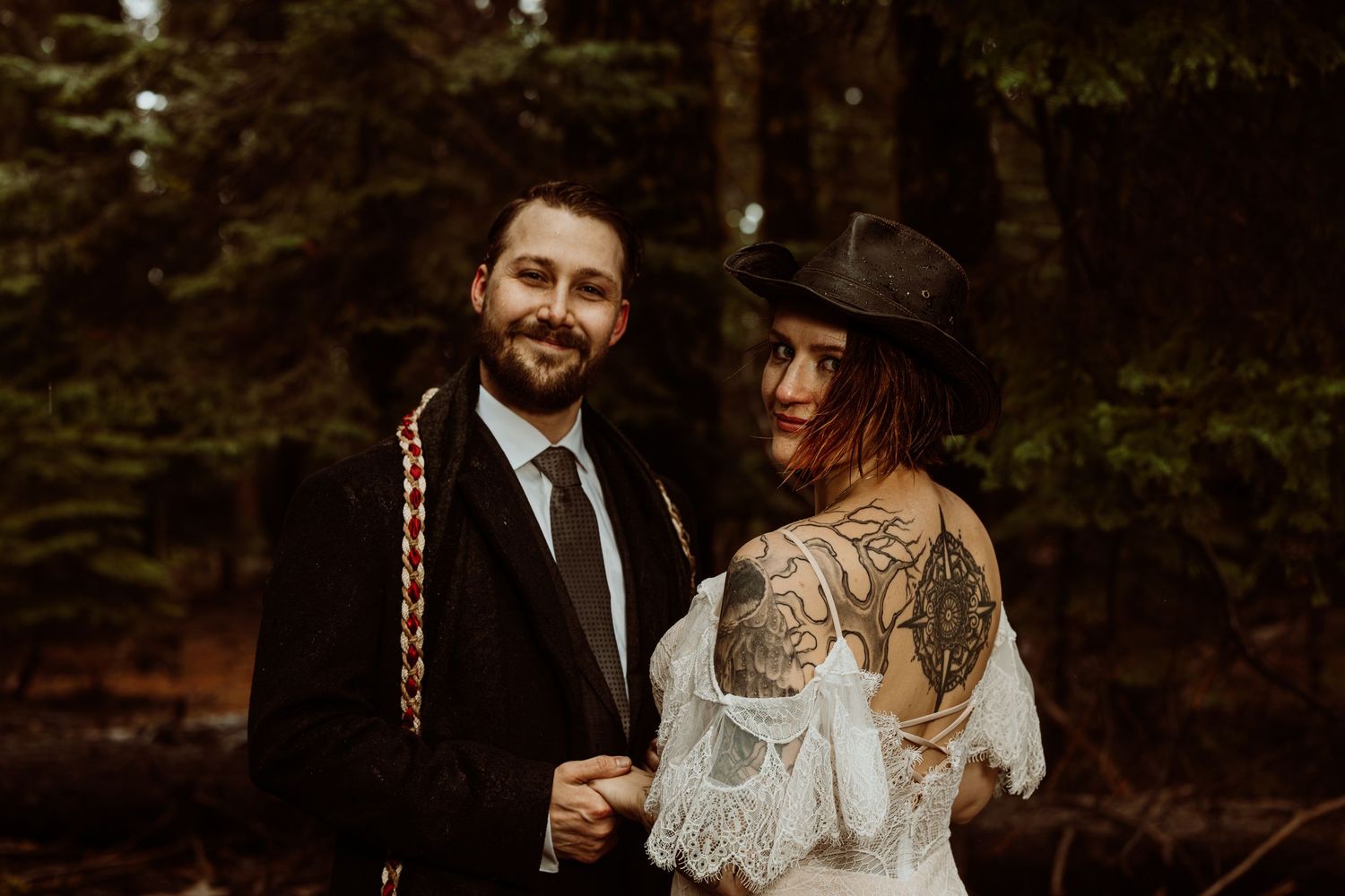 Couple in wedding attire poses in forest, groom in black suit with tie, bride in lace dress and cowboy hat with tattoos.