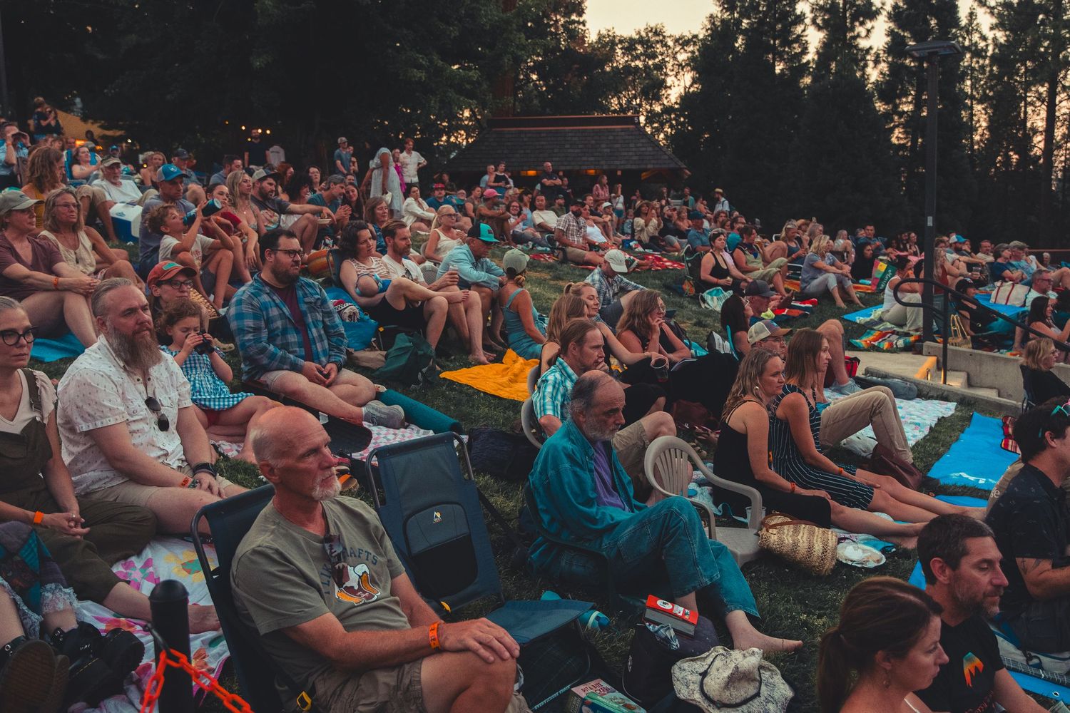 Large outdoor concert audience seated on chairs and blankets at dusk surrounded by tall trees.