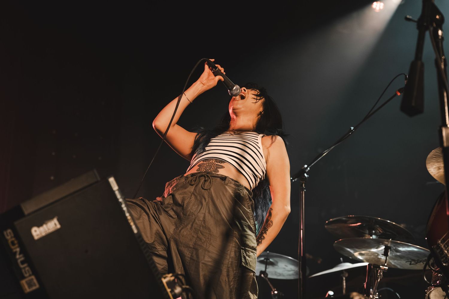 Female vocalist sings passionately into microphone on dark stage wearing striped top and baggy pants.