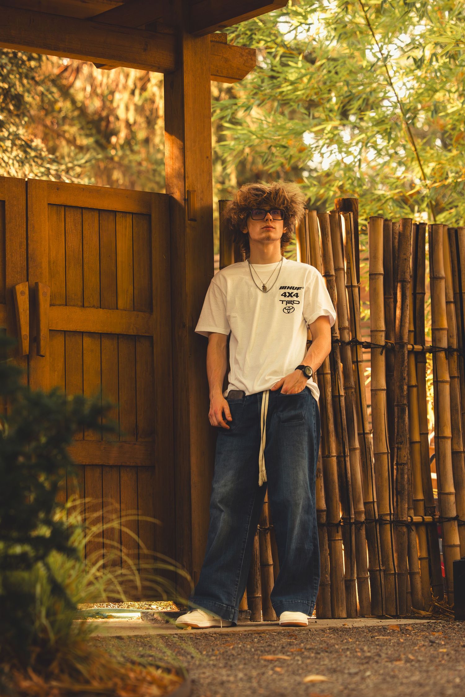 Curly-haired teen in white graphic tee and wide-leg jeans posing by bamboo fence in garden.