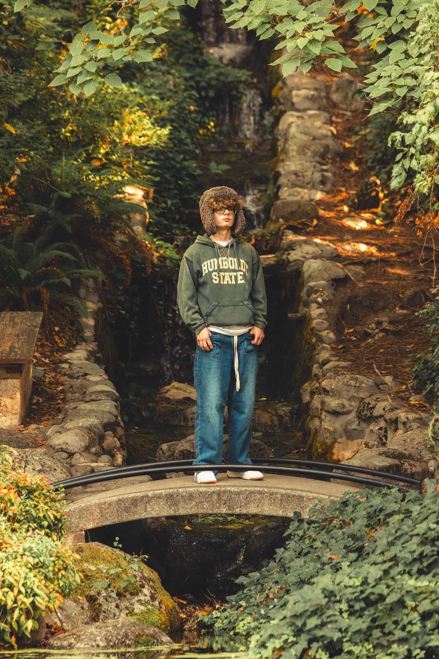 A young man in a Humboldt State hoodie stands on a small bridge in a lush garden.