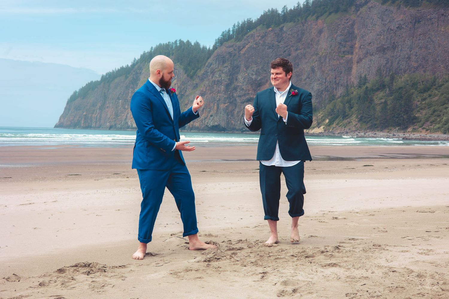Two grooms in blue suits dancing barefoot on a beach with rocky cliffs in the background.