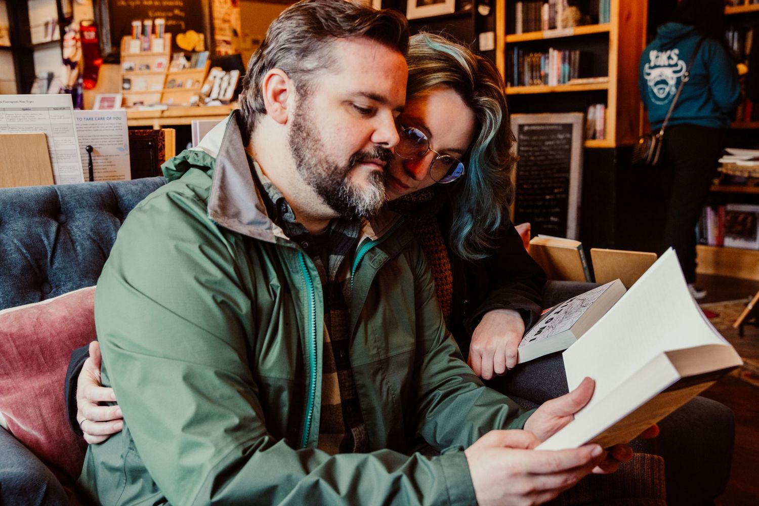 A bearded man reading a book while a blue-haired woman snuggles close in a bookstore.