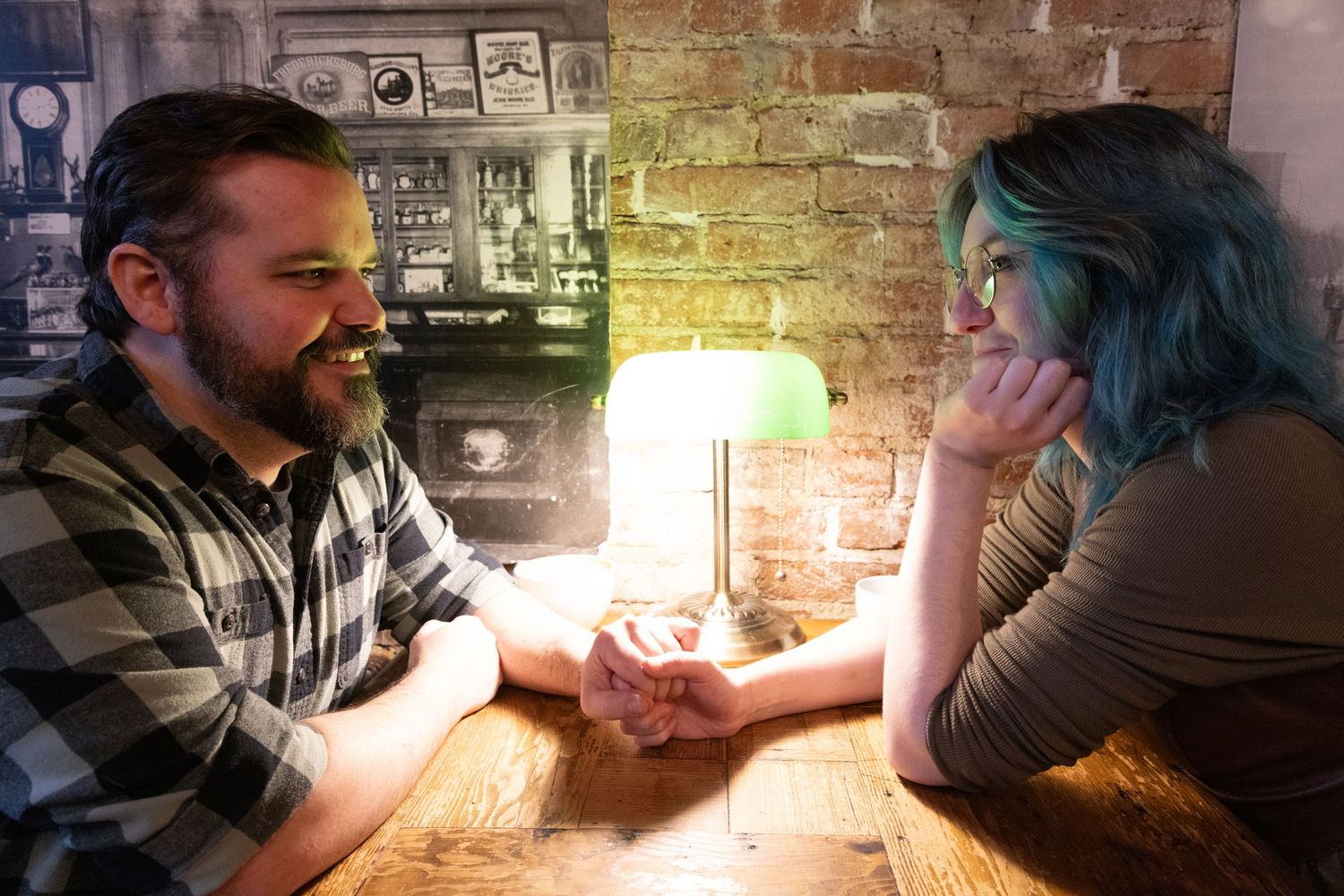Couple holding hands across a wooden table by a green lamp in a cozy brick-walled bar.