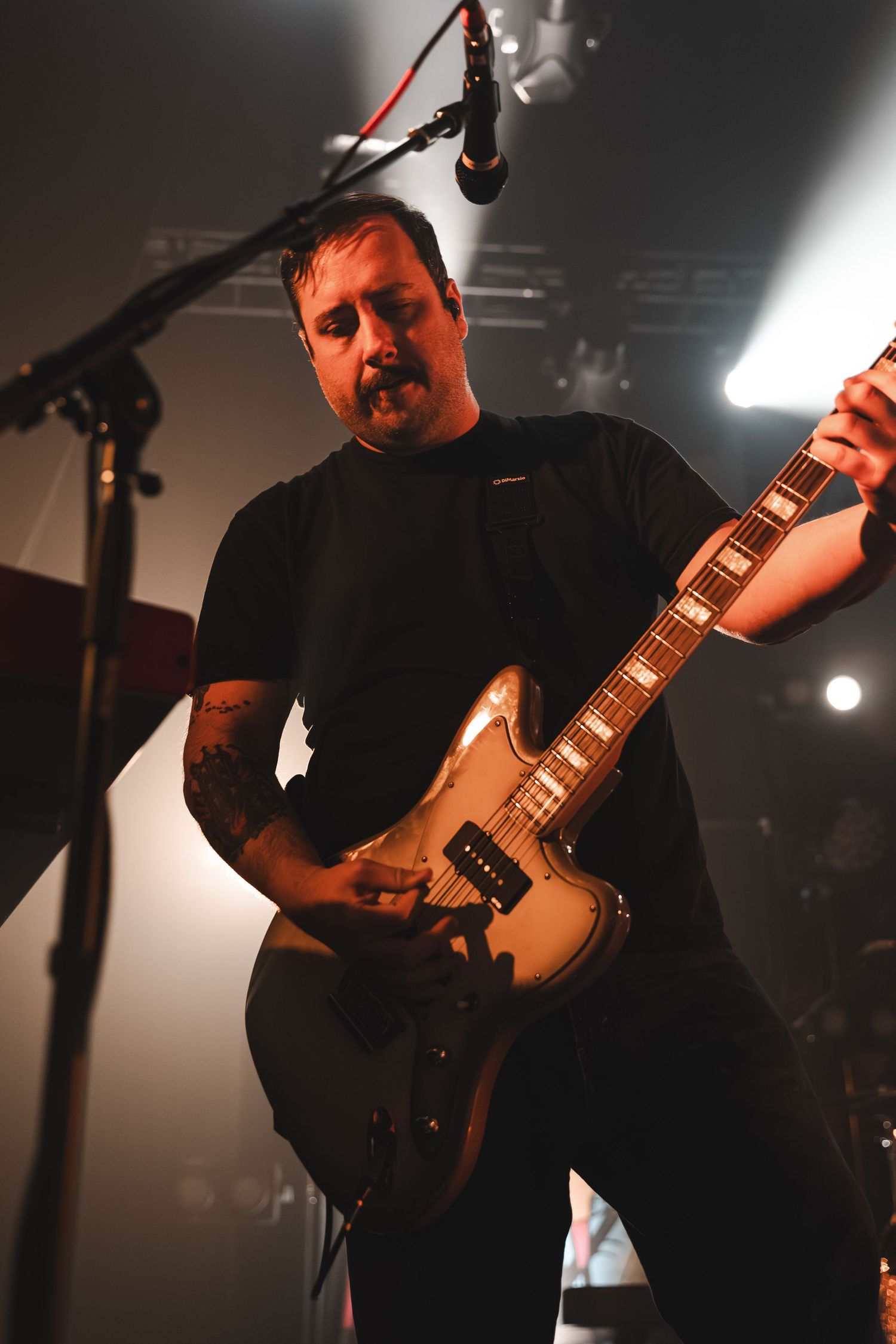 Tattooed guitarist plays white electric guitar under stage lights at a live rock concert.