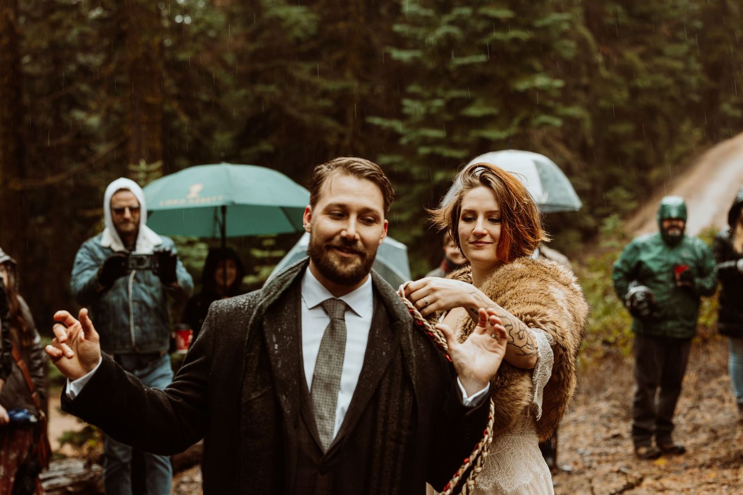 Groom gestures expressively beside bride in fur wrap during rainy forest wedding ceremony.