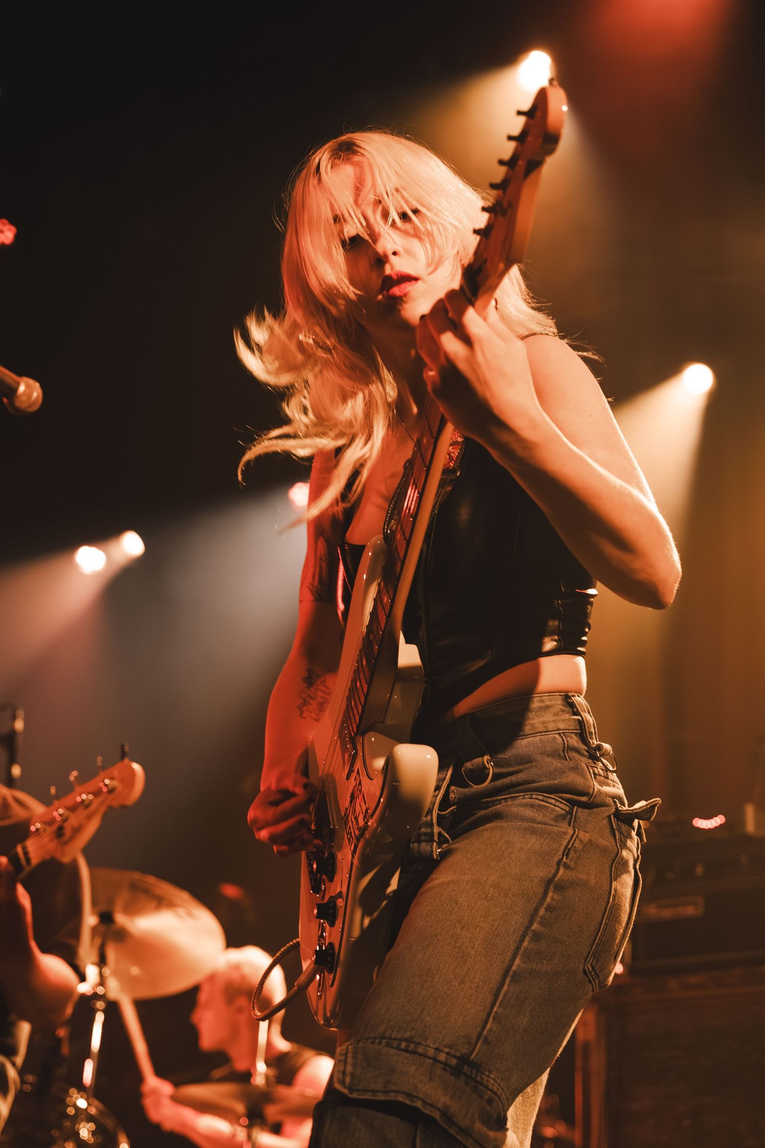 Energetic blonde female guitarist headbanging while playing electric guitar under warm concert stage lighting.