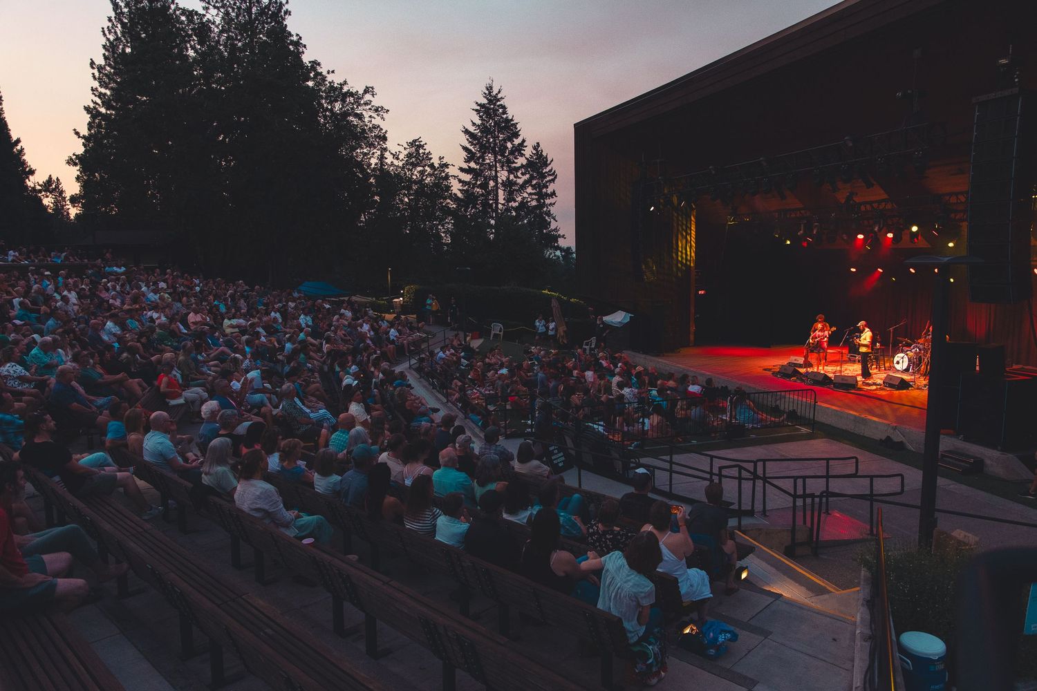 Packed outdoor amphitheater audience watching a twilight concert performance on a warmly lit stage.