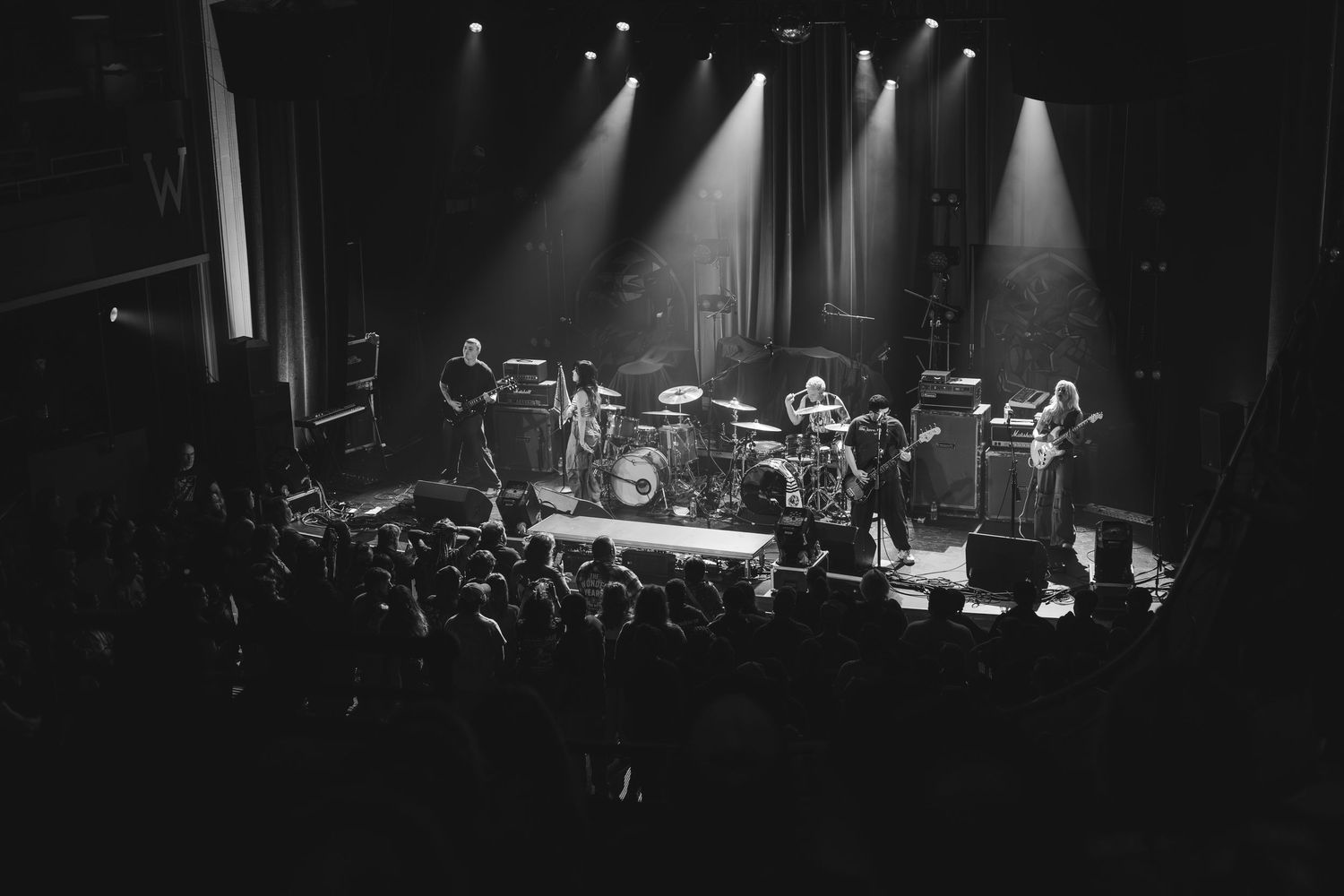 Black and white wide shot of full band performing on stage before a packed concert crowd.