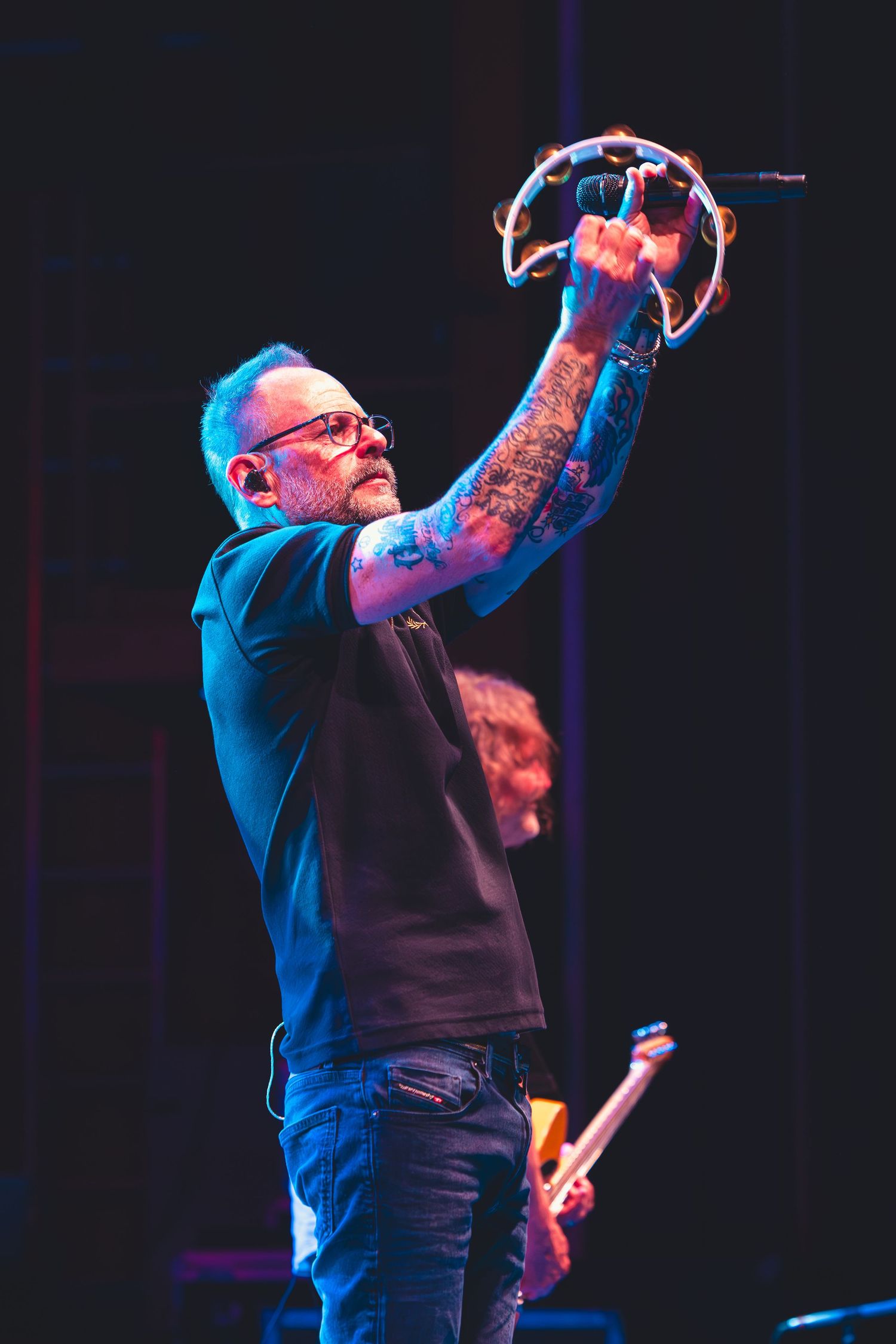 Tattooed performer raises a tambourine overhead on a darkened concert stage.