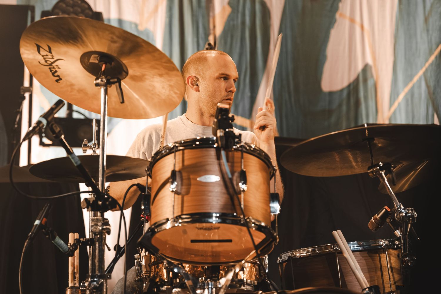 Bald drummer playing a wooden drum kit with Zildjian cymbals on a live concert stage.