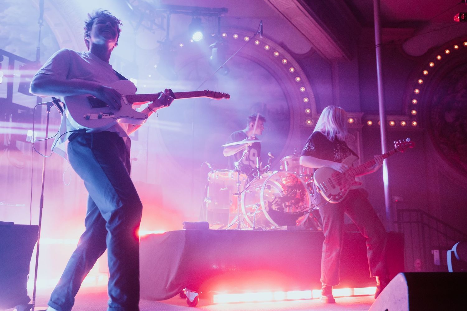 Three-piece band performing on stage with atmospheric purple and pink lighting and fog effects.