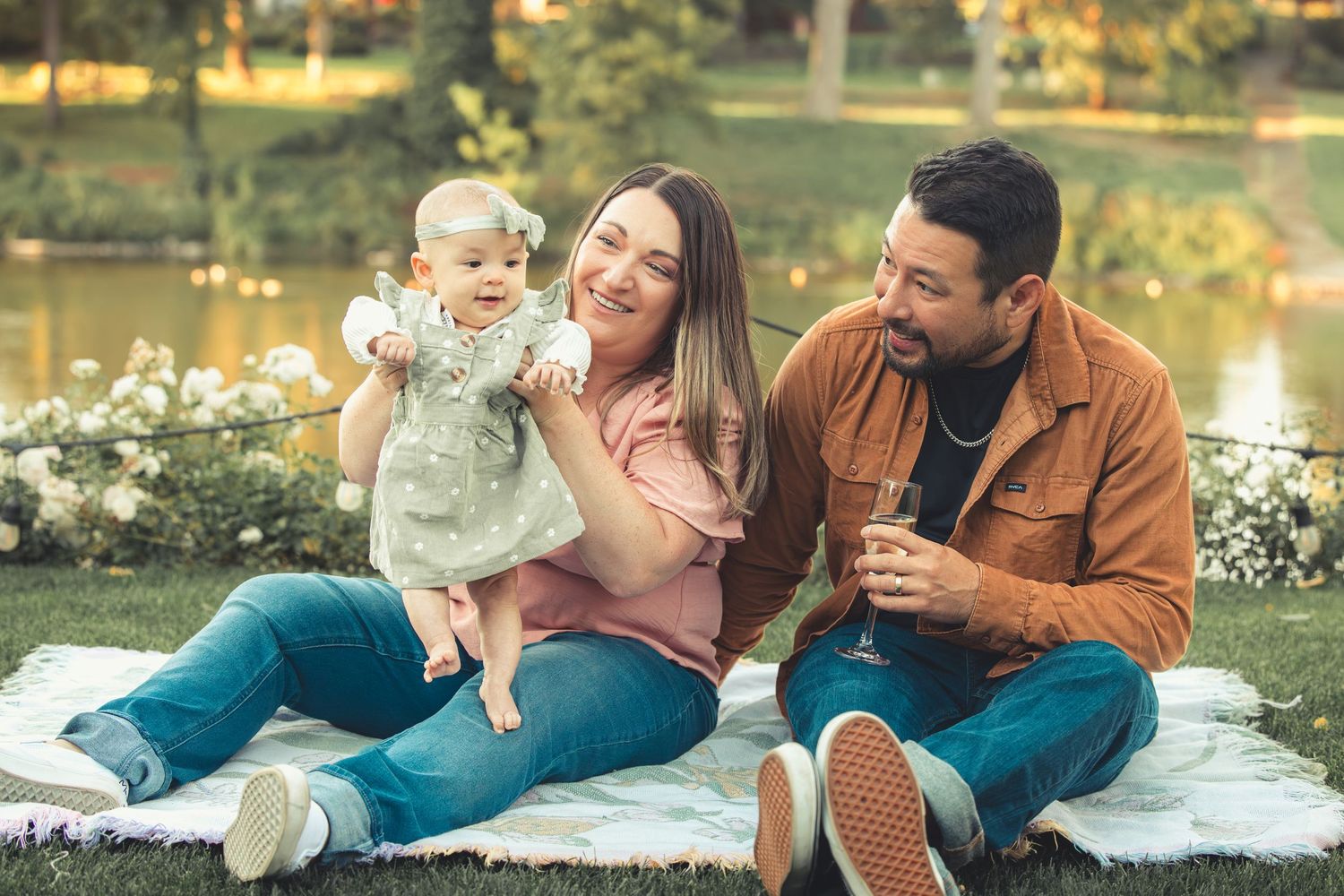 Parents sitting on a blanket in a park admiring their adorable baby girl.
