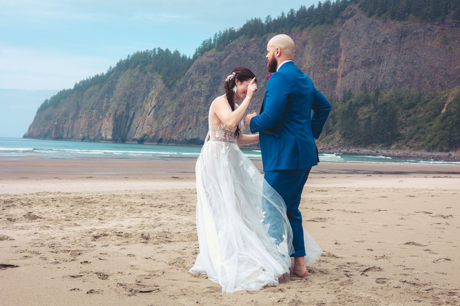 Groom in blue suit laughs with bride in white gown on sandy beach, forested rocky cliffs in background.
