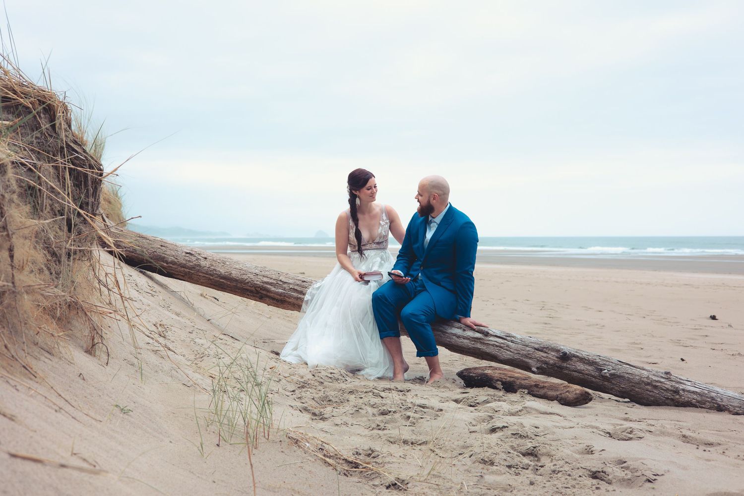 Bride in white gown and groom in blue suit sit closely on driftwood, smiling at each other on a beach.