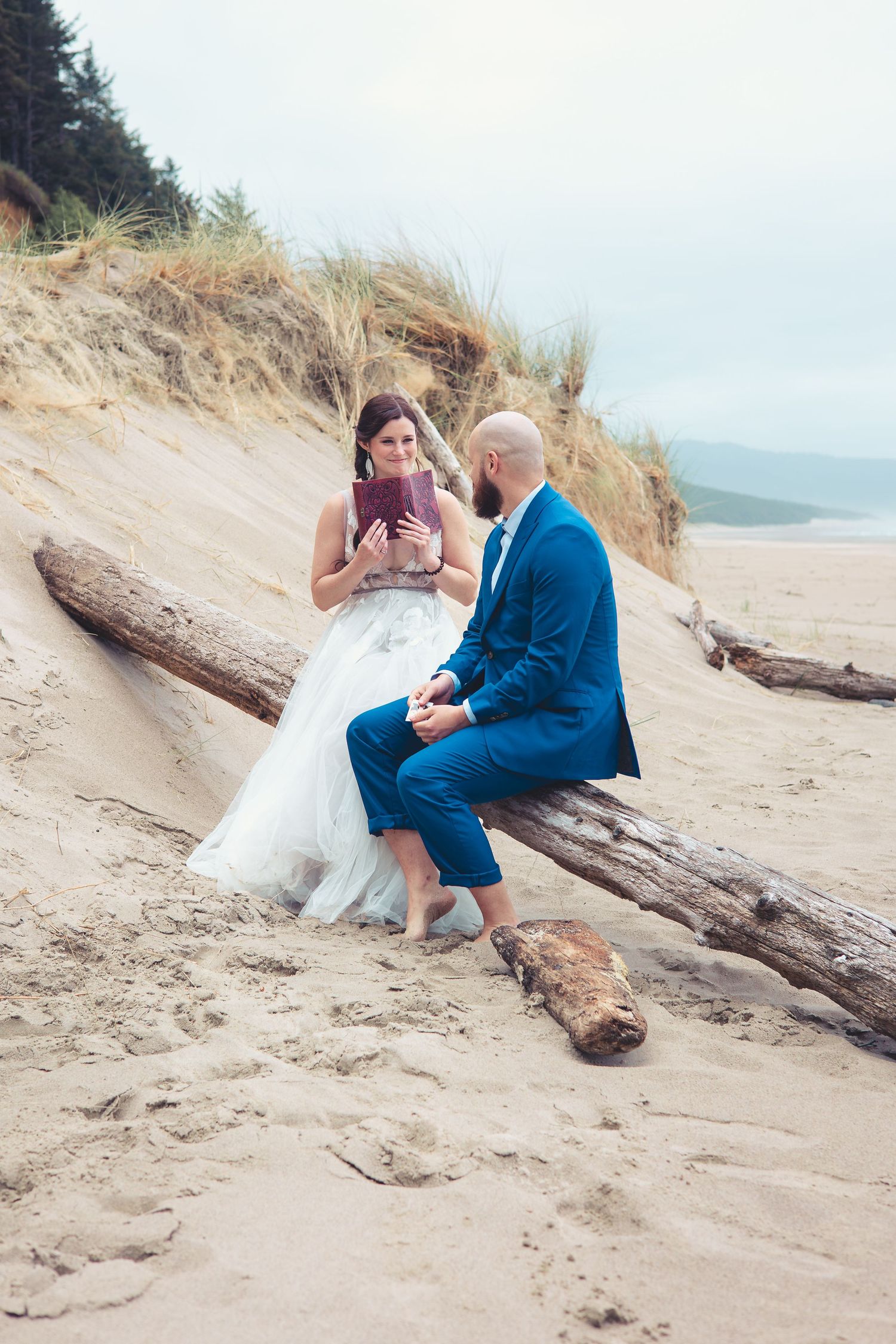 Bride reading vows from a purple book to groom in blue suit seated on driftwood at a sandy beach.