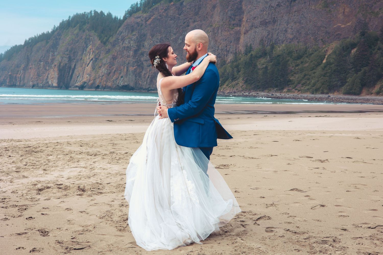Bride and groom embrace on sandy beach, groom lifting bride slightly, dramatic rocky cliffs behind them.
