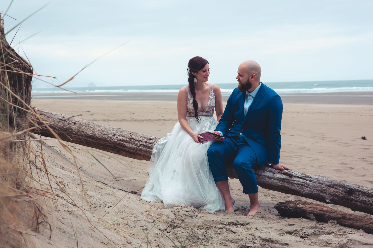 Bride in white dress and bald groom in blue suit sit on driftwood gazing at each other on a beach.