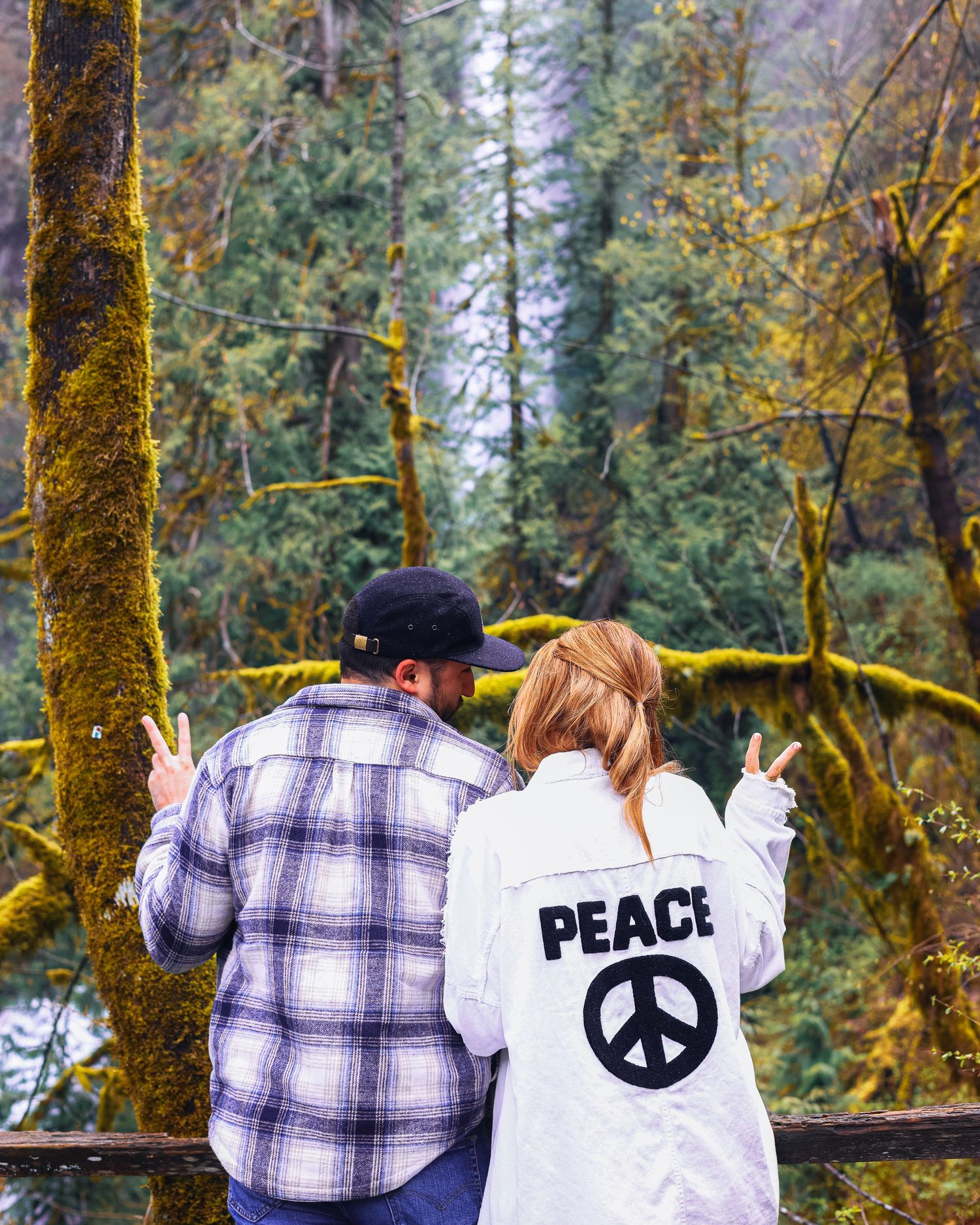 A couple makes peace signs while admiring a moss-covered forest landscape.