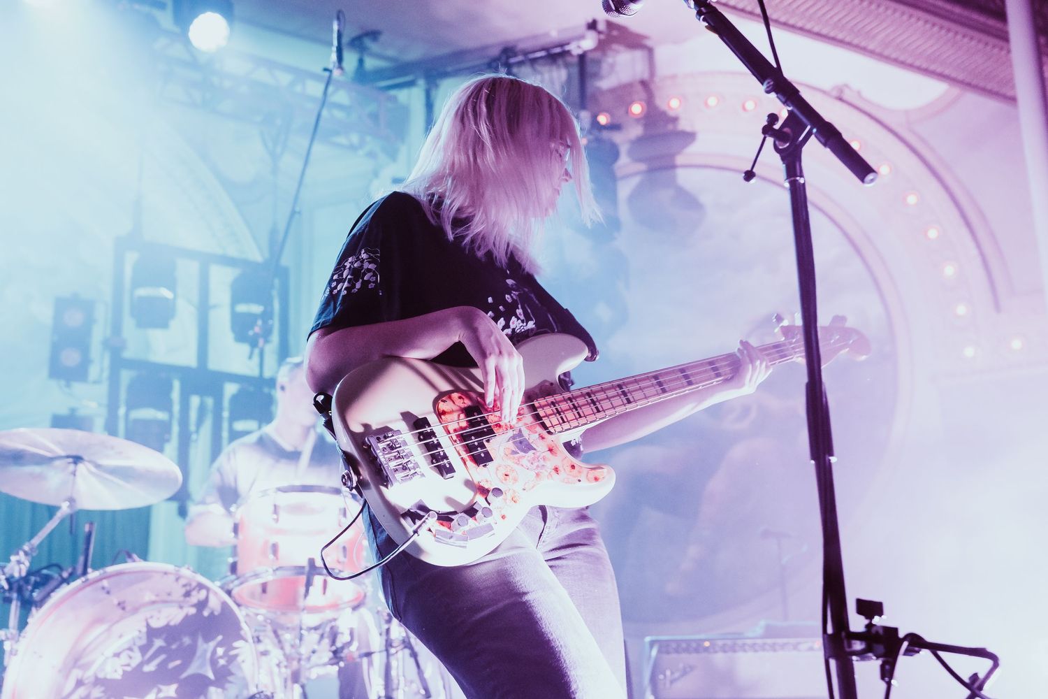 Blonde female bassist playing on stage under vibrant pink and blue concert lighting.
