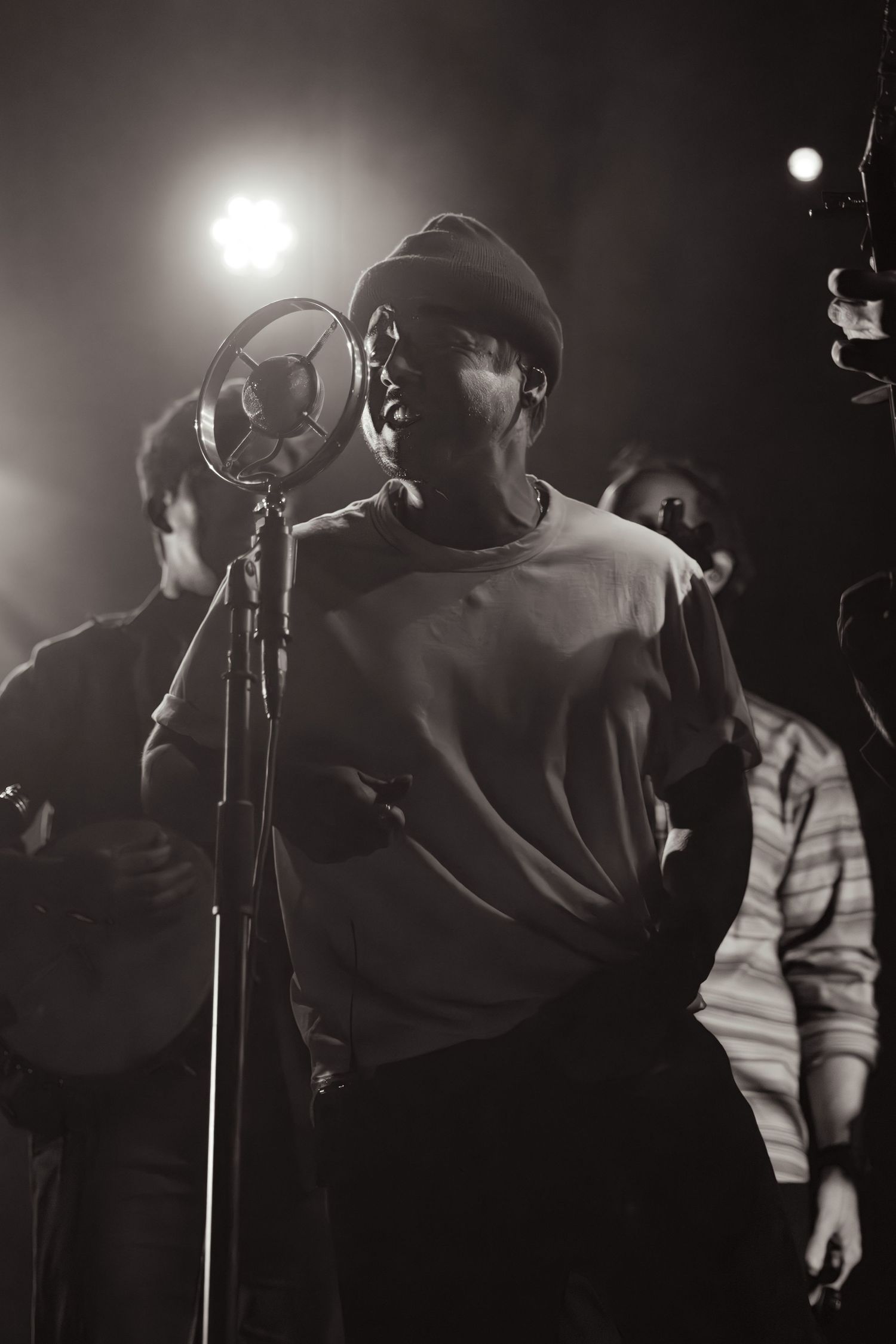 Black and white shot of a male vocalist singing into a studio microphone under bright stage lights.