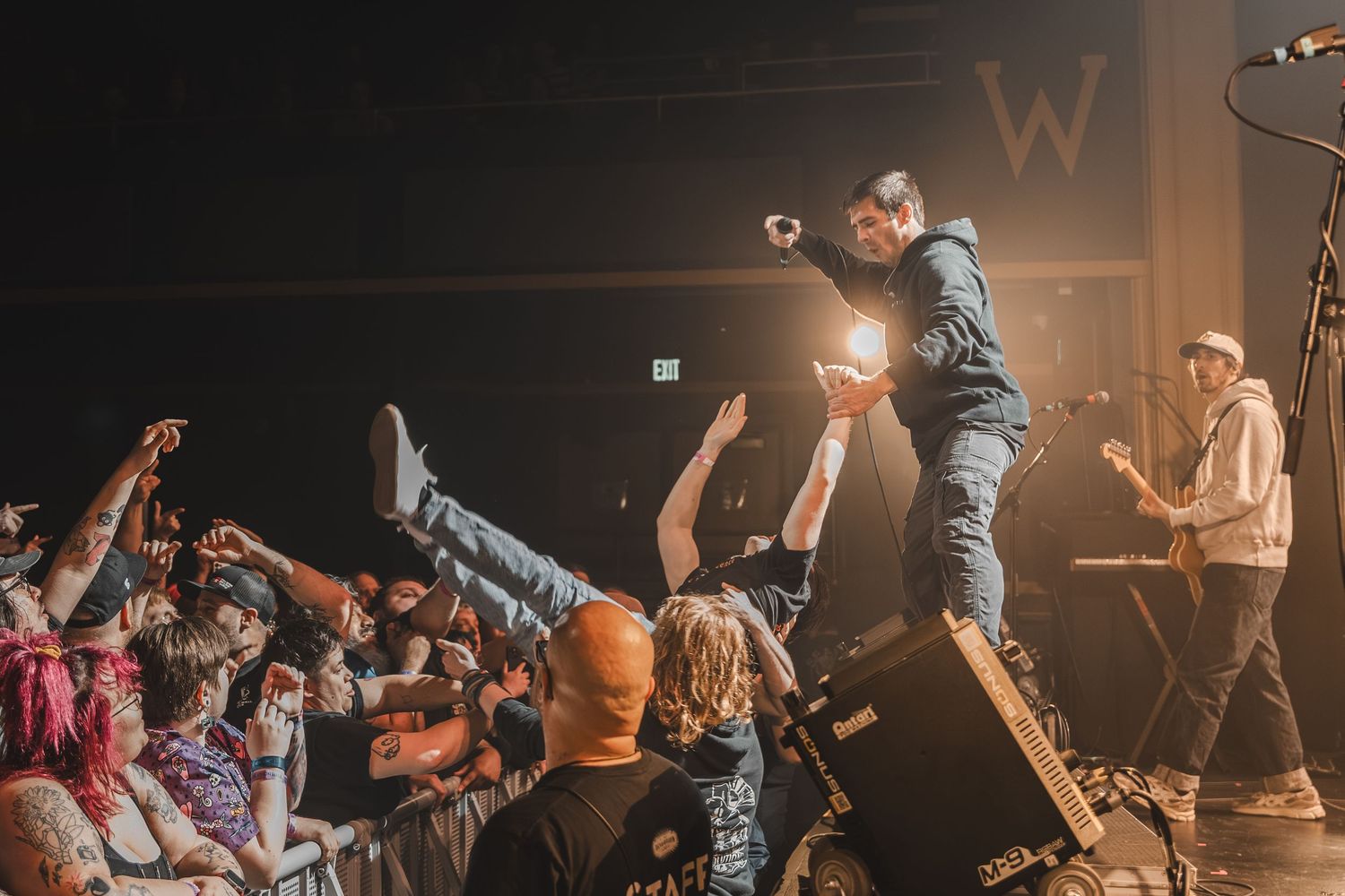 Energetic performer stands on speaker interacting with crowd of fans with hands raised at live concert.