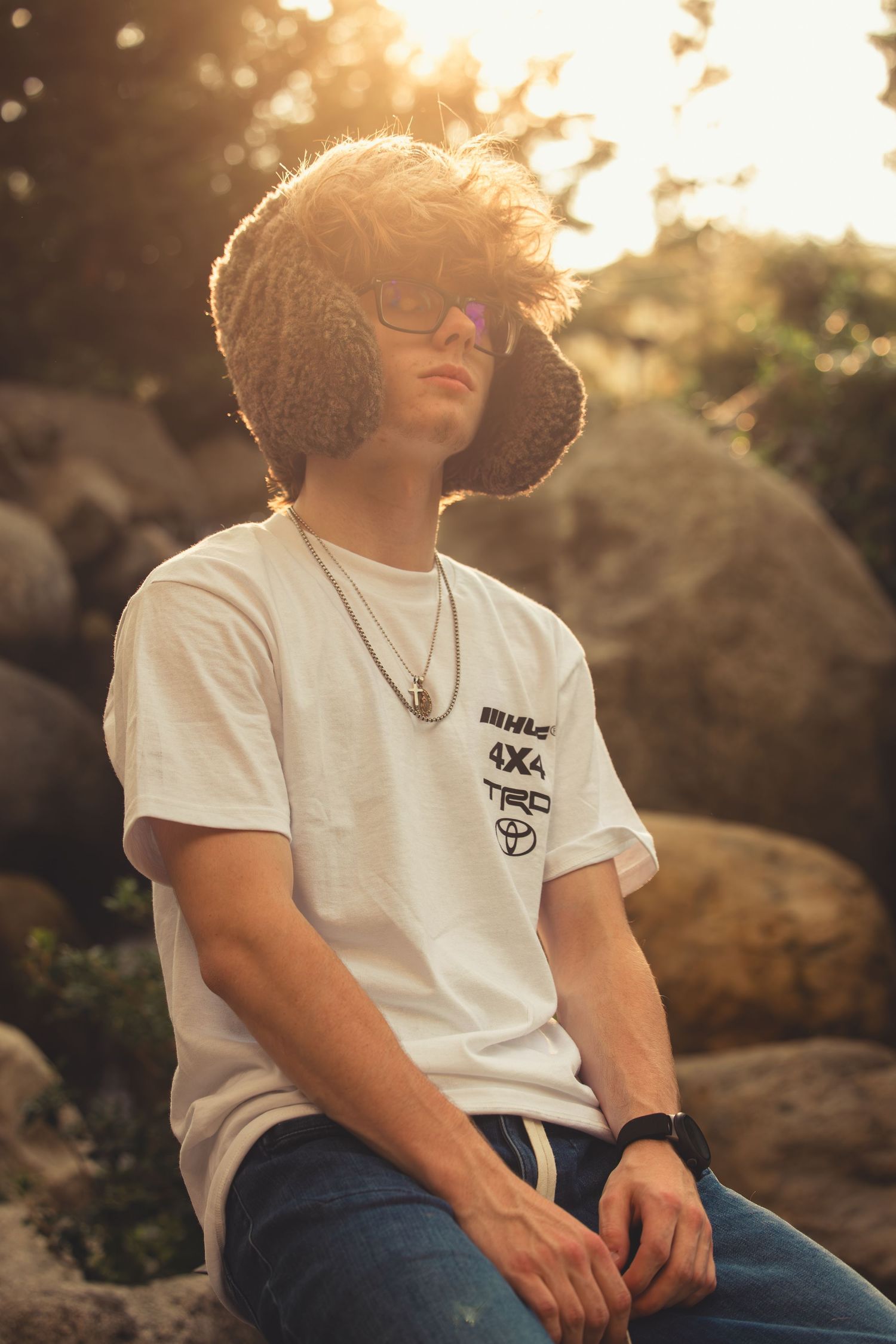Teen boy in white 4x4 TRD t-shirt and jeans sitting on rocks wearing a bucket hat at sunset.