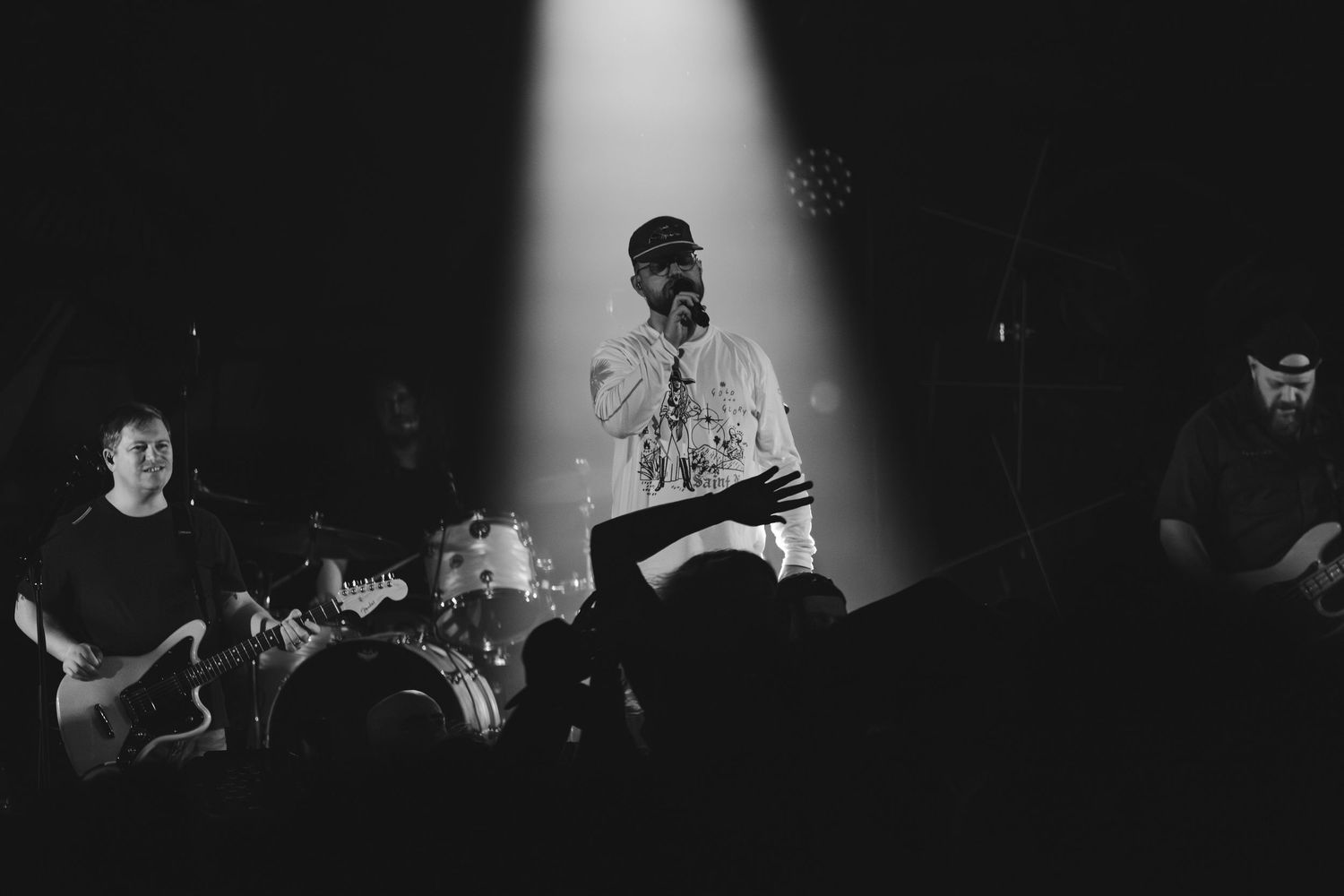 Black and white spotlight photo of vocalist performing on stage with band members behind.