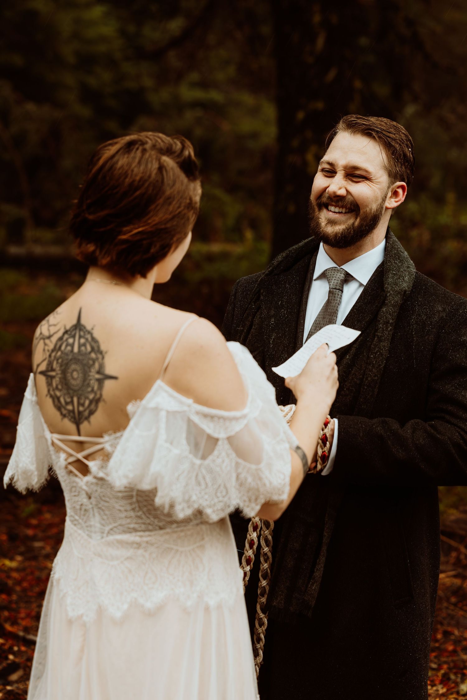 Groom laughs joyfully while reading vows to tattooed bride in a woodland wedding ceremony.