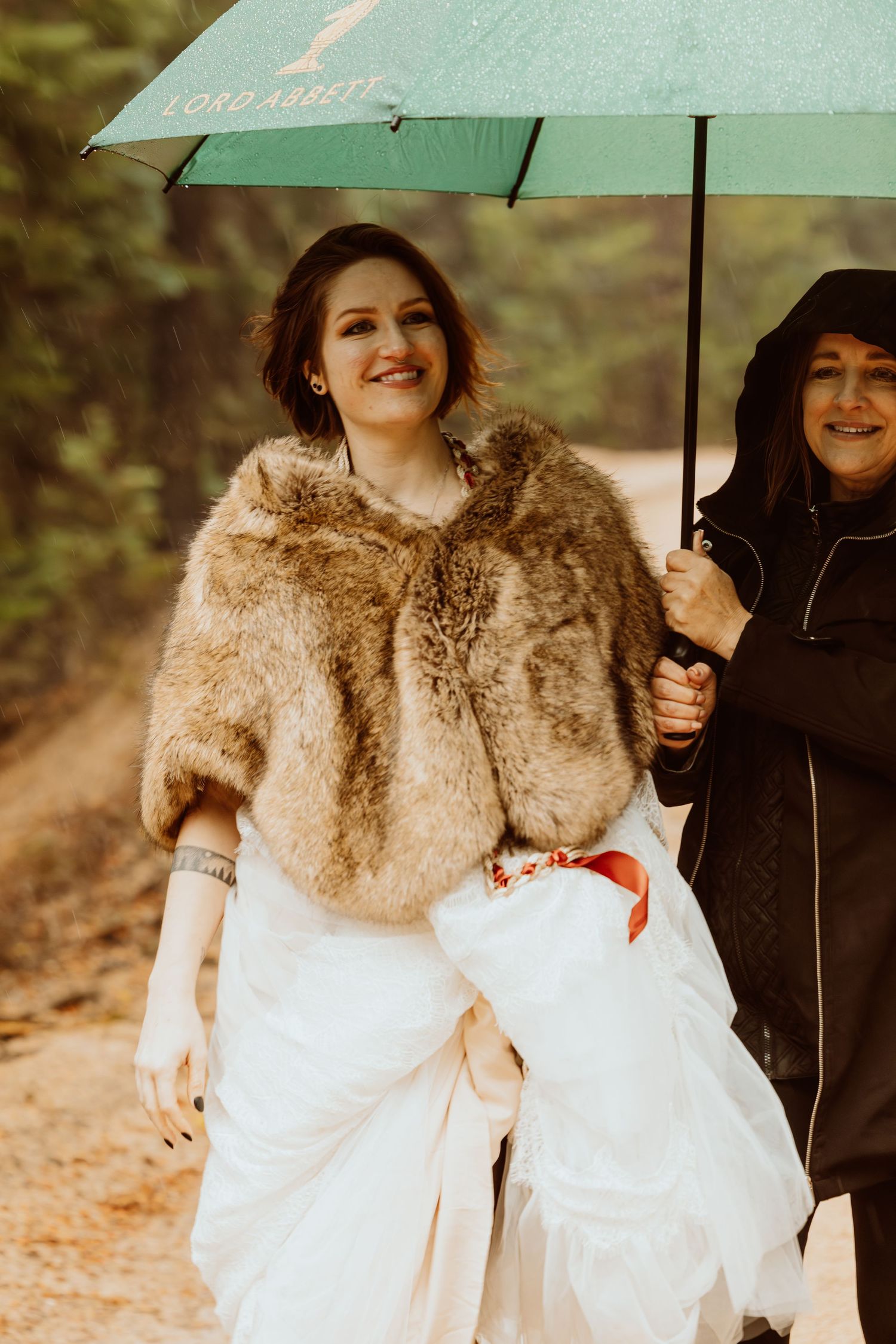 Smiling bride in fur stole walks under green Lord Ashley umbrella held by woman in black jacket.