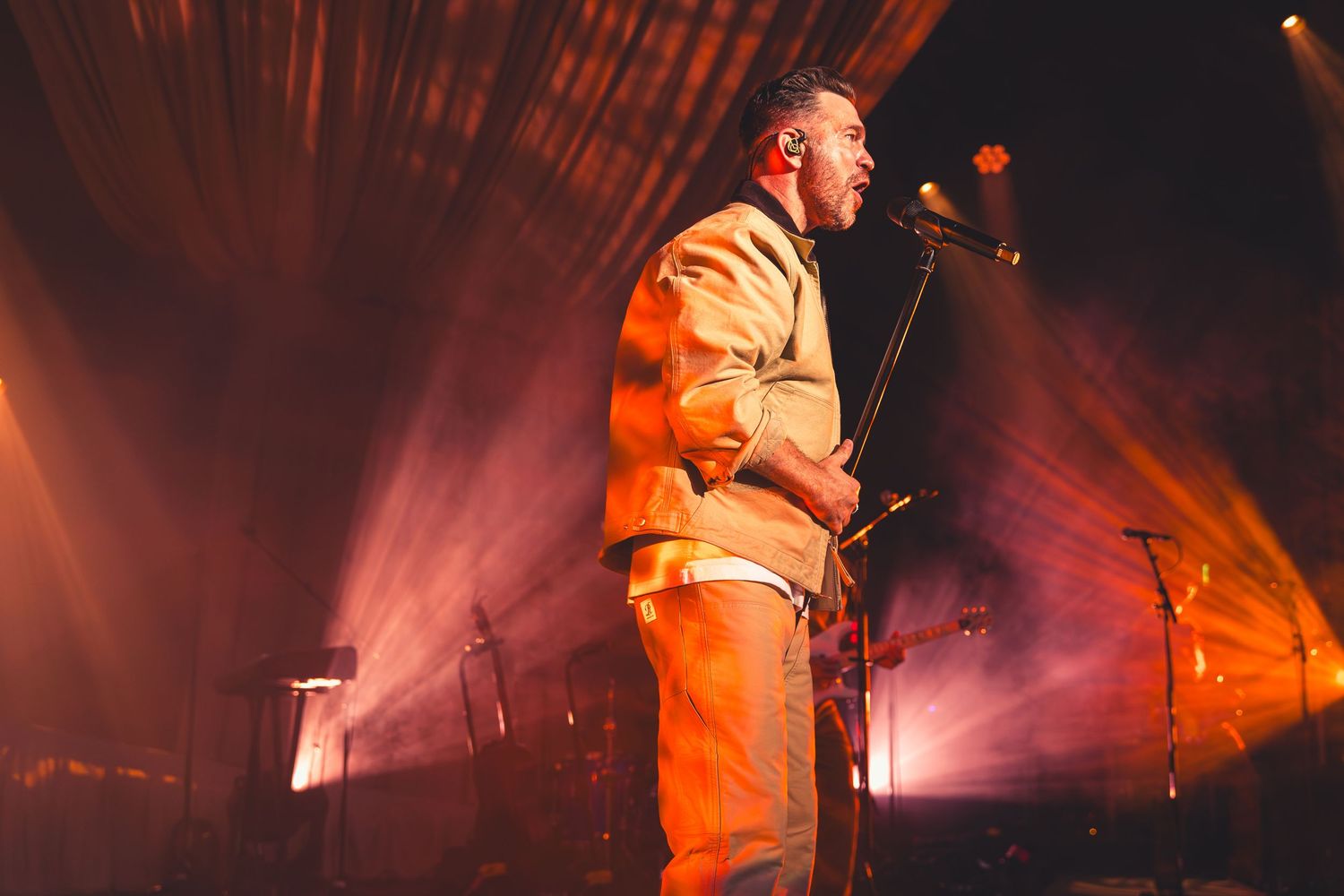 A male singer performs on stage in an orange outfit under warm pink spotlights.