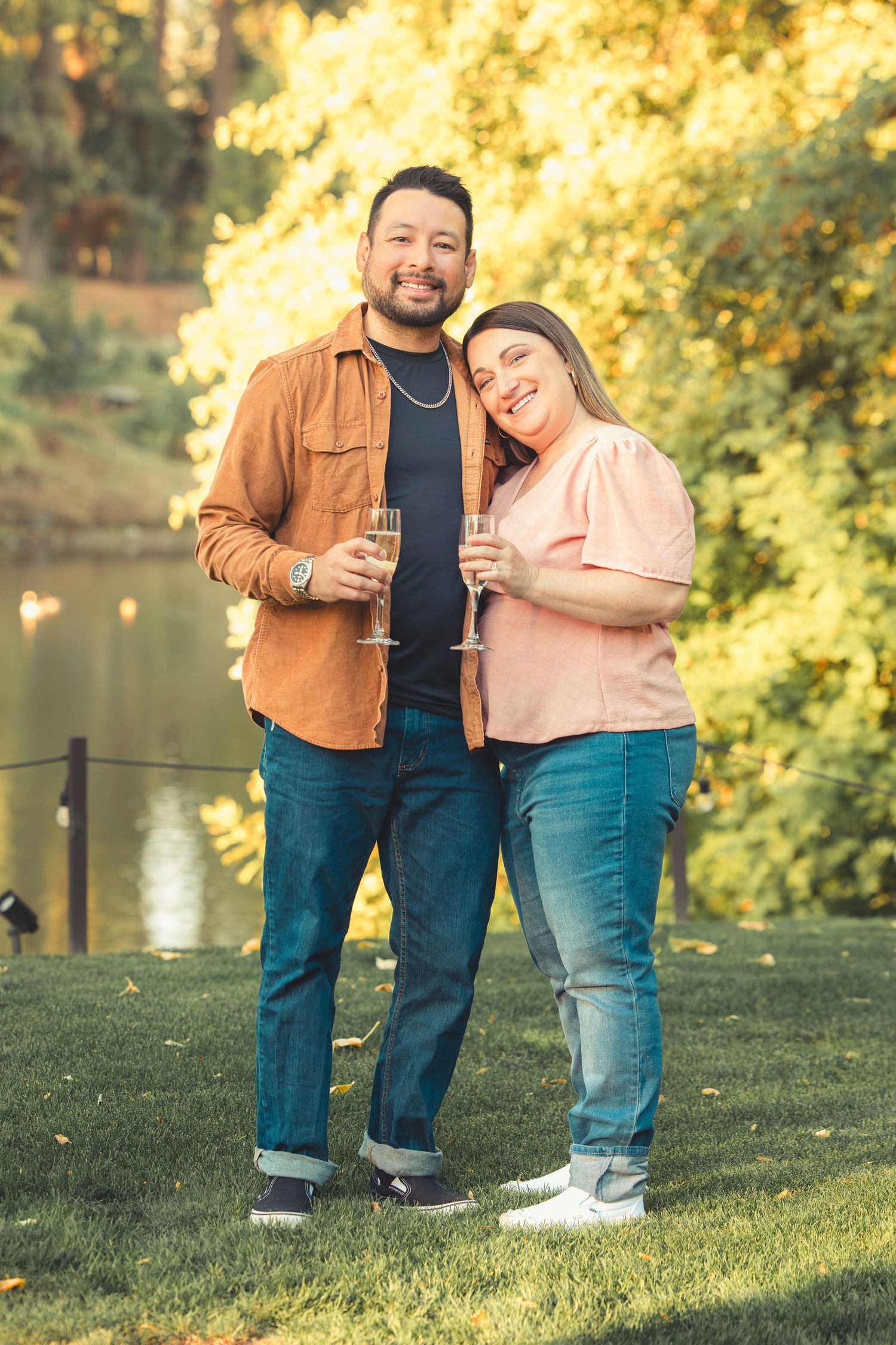 Smiling couple cuddling with champagne glasses in a sunlit park with vibrant fall foliage backdrop.
