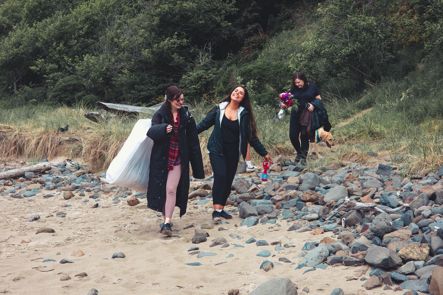 Group of women walking on a rocky beach carrying a white dress and colorful bouquet outdoors.