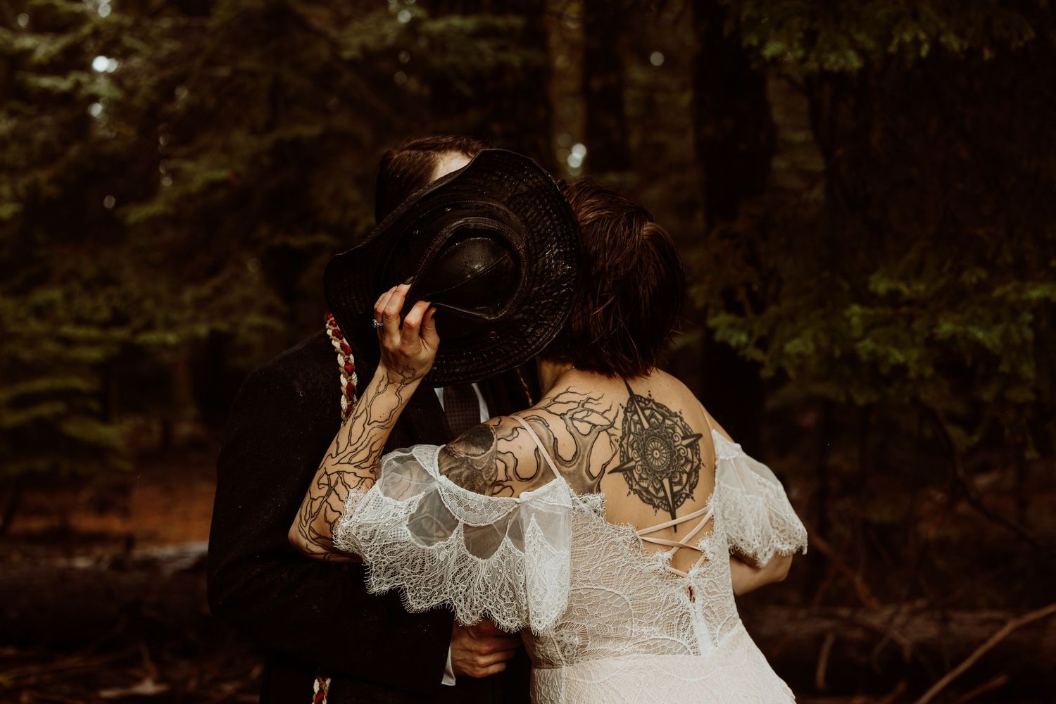Tattooed bride in white lace dress and groom share a kiss hidden behind her black cowboy hat in a forest setting.