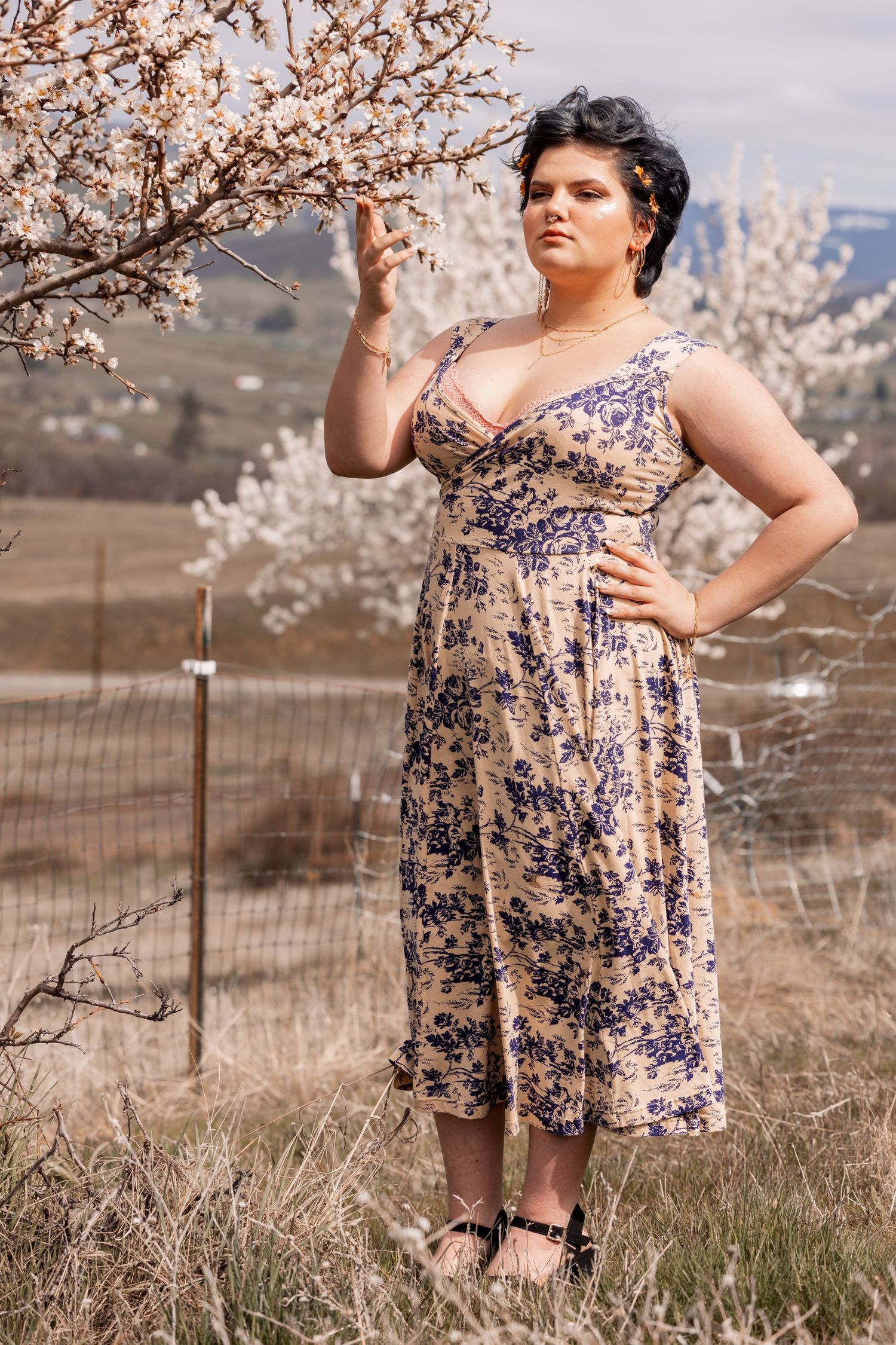 Woman in blue floral midi dress standing near blooming tree, touching white blossoms.