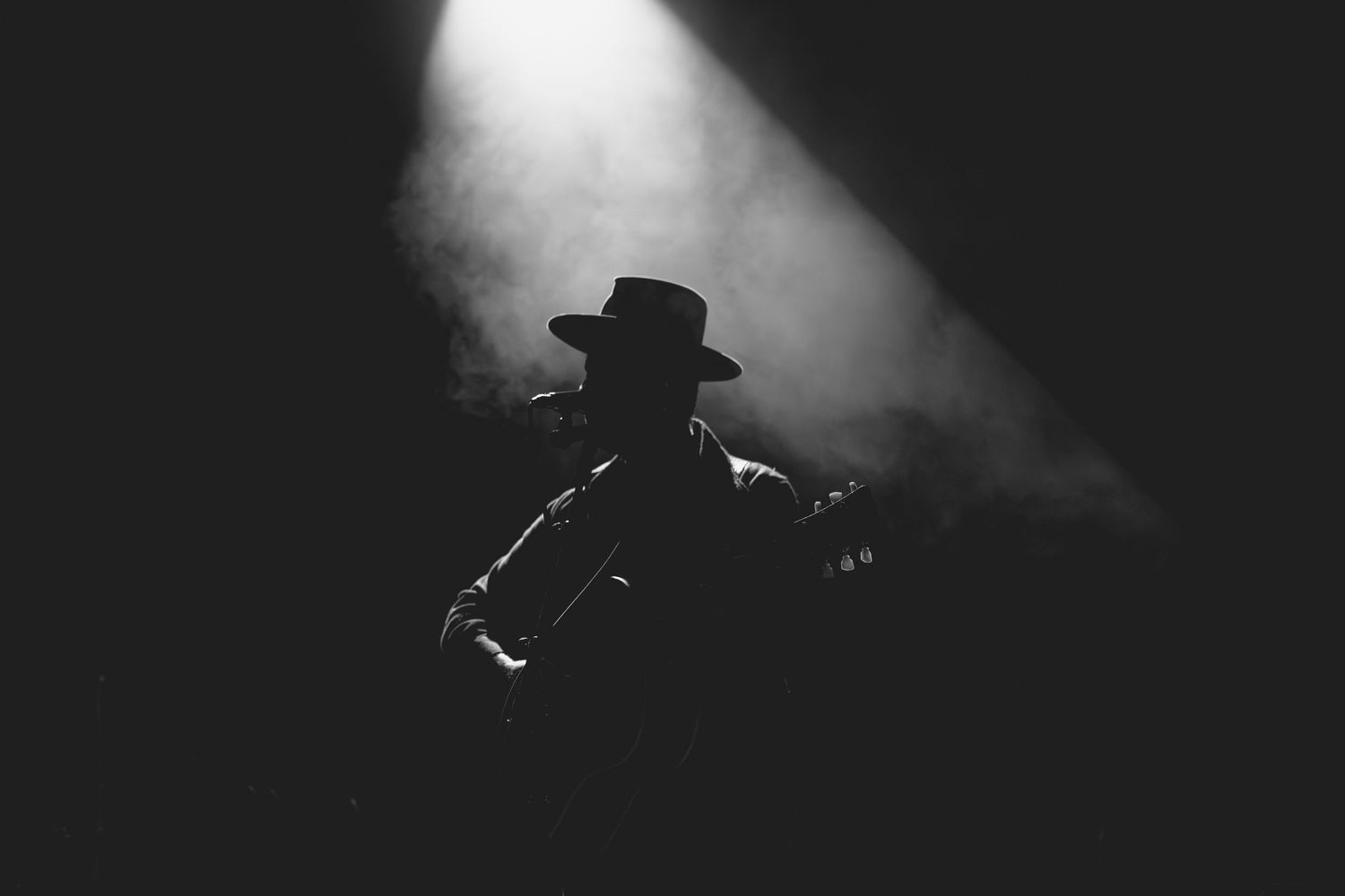 Black and white silhouette of guitarist wearing cowboy hat performing on dark smoky concert stage.