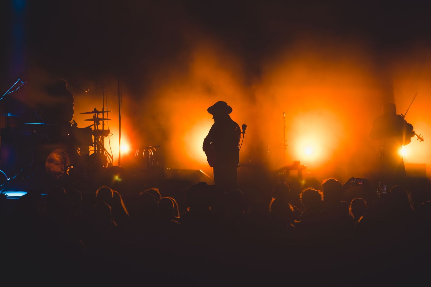 Silhouetted guitarist in cowboy hat performing before crowd with dramatic orange and amber fog-filled stage lighting.