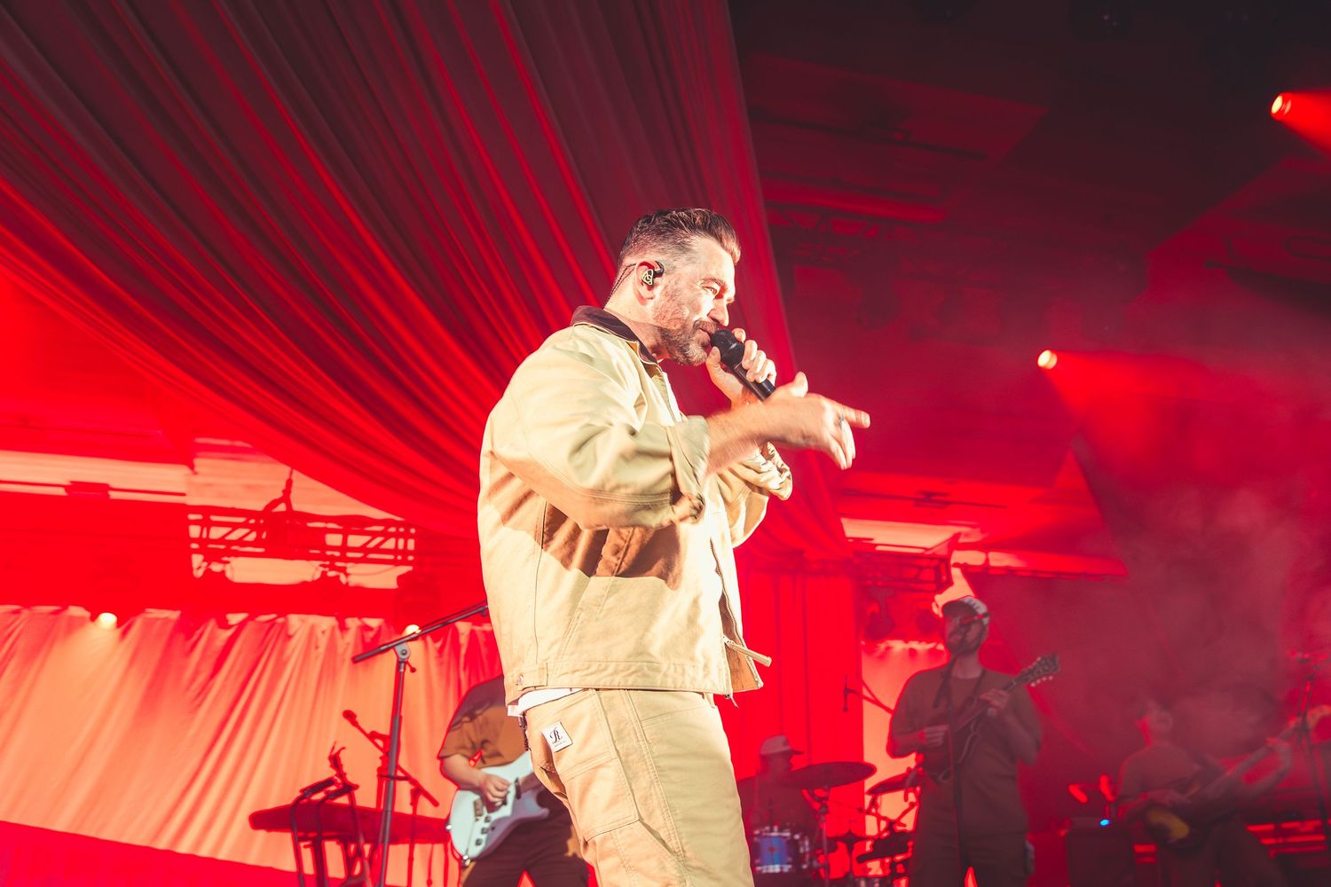 A male performer in a beige outfit sings into a microphone under dramatic red stage lighting.