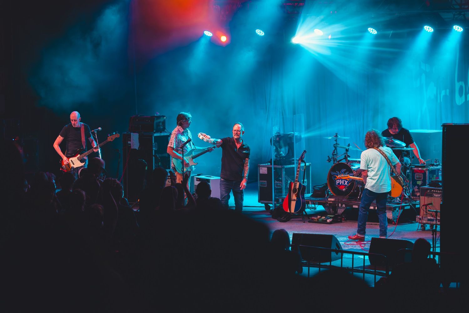 Five-piece band performing on a darkened stage with teal and red concert lighting during a live show.