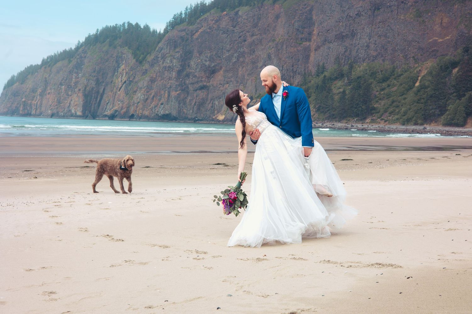 Couple sharing a kiss on the beach with a dog nearby and scenic coastal cliffs in the background.