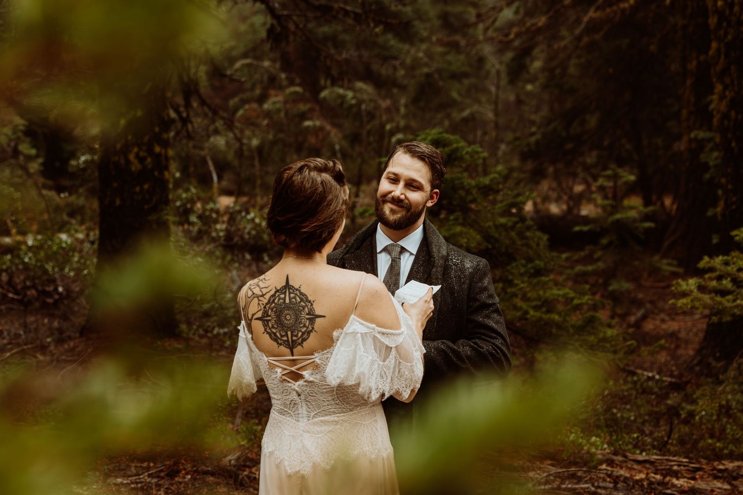 Groom smiling at bride with intricate back tattoo in a lush forest wedding setting.