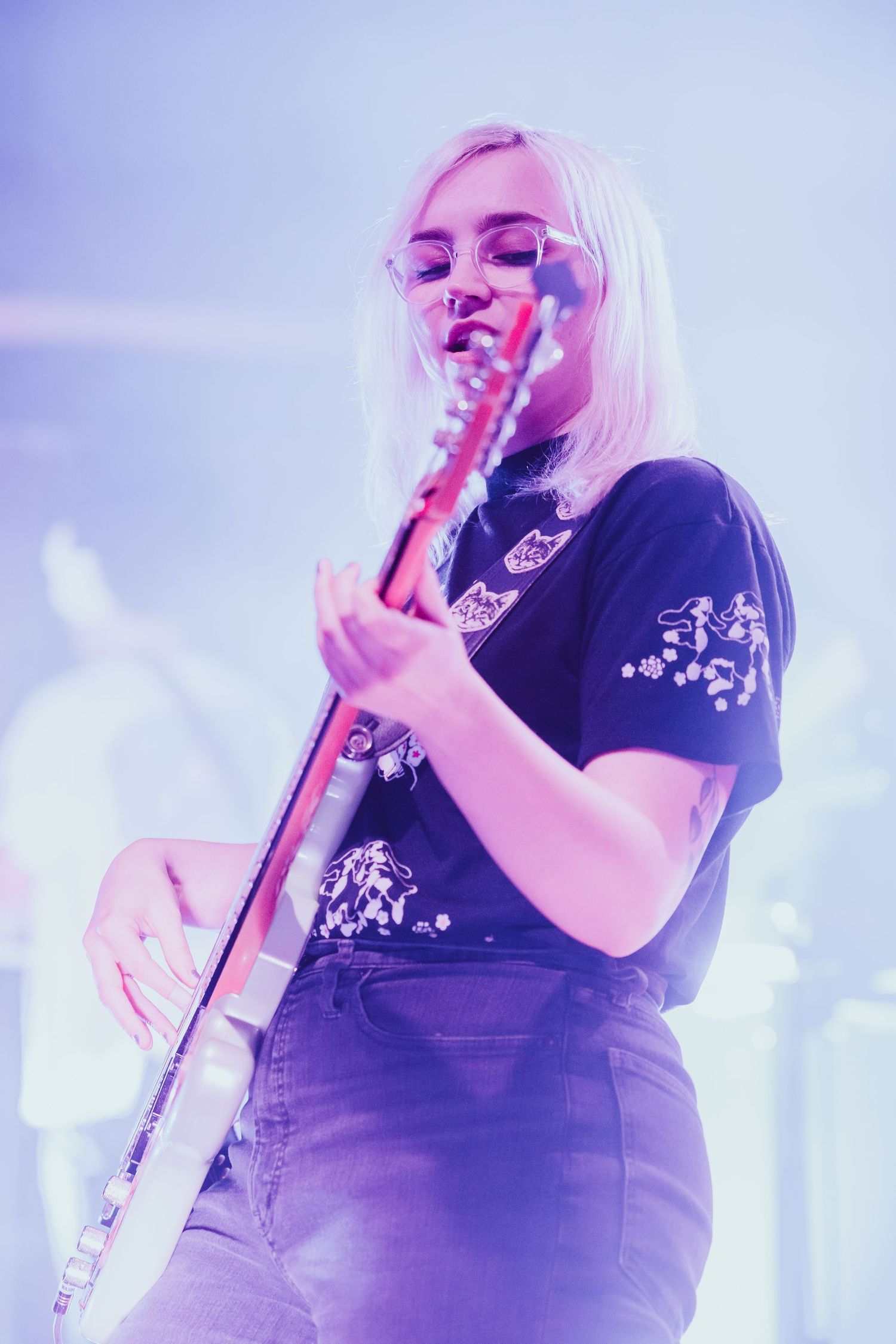 Blonde female guitarist performs on stage bathed in purple concert lighting.