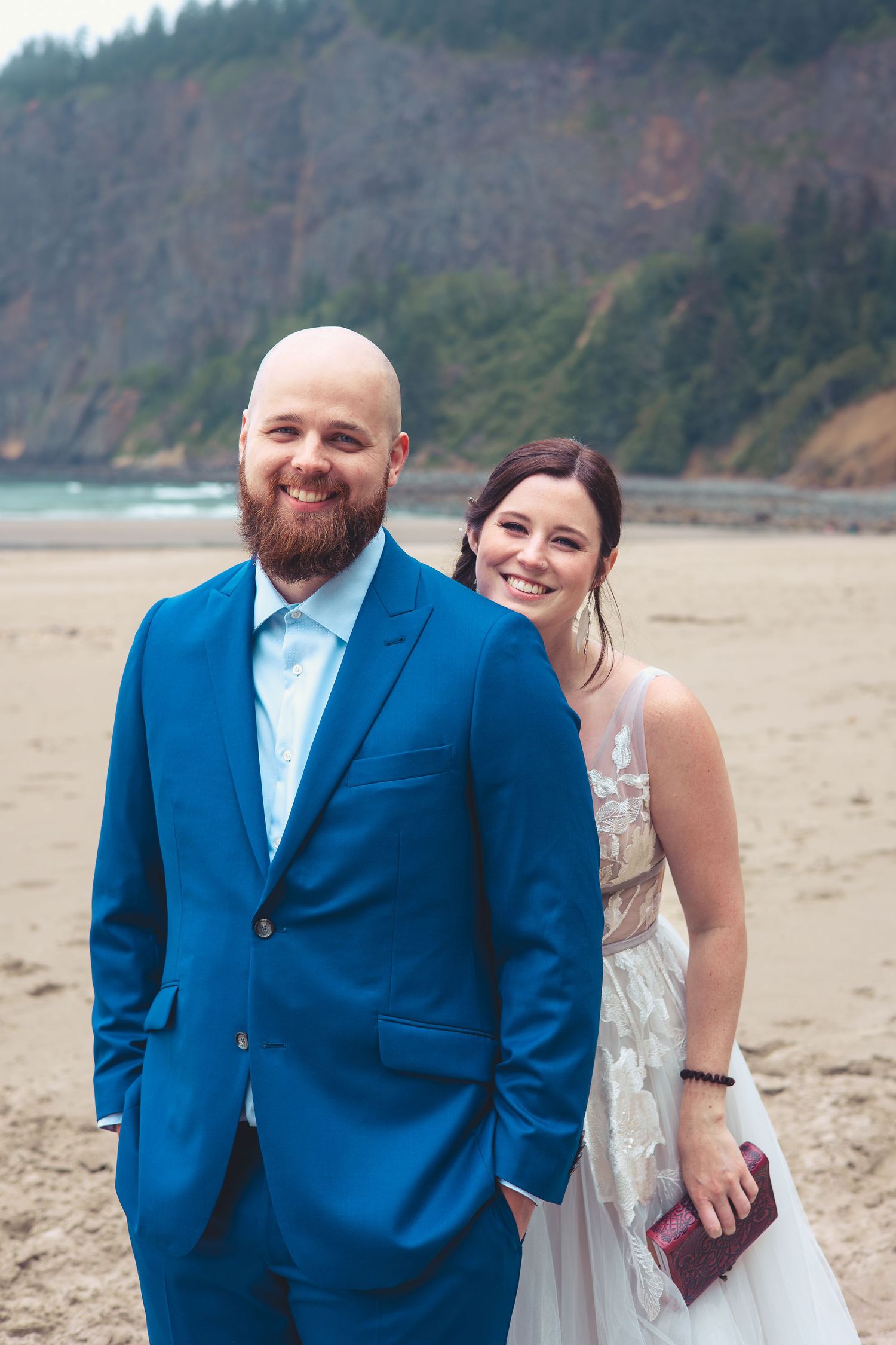 Smiling couple in wedding attire pose on a beach with dramatic rocky cliffs behind.