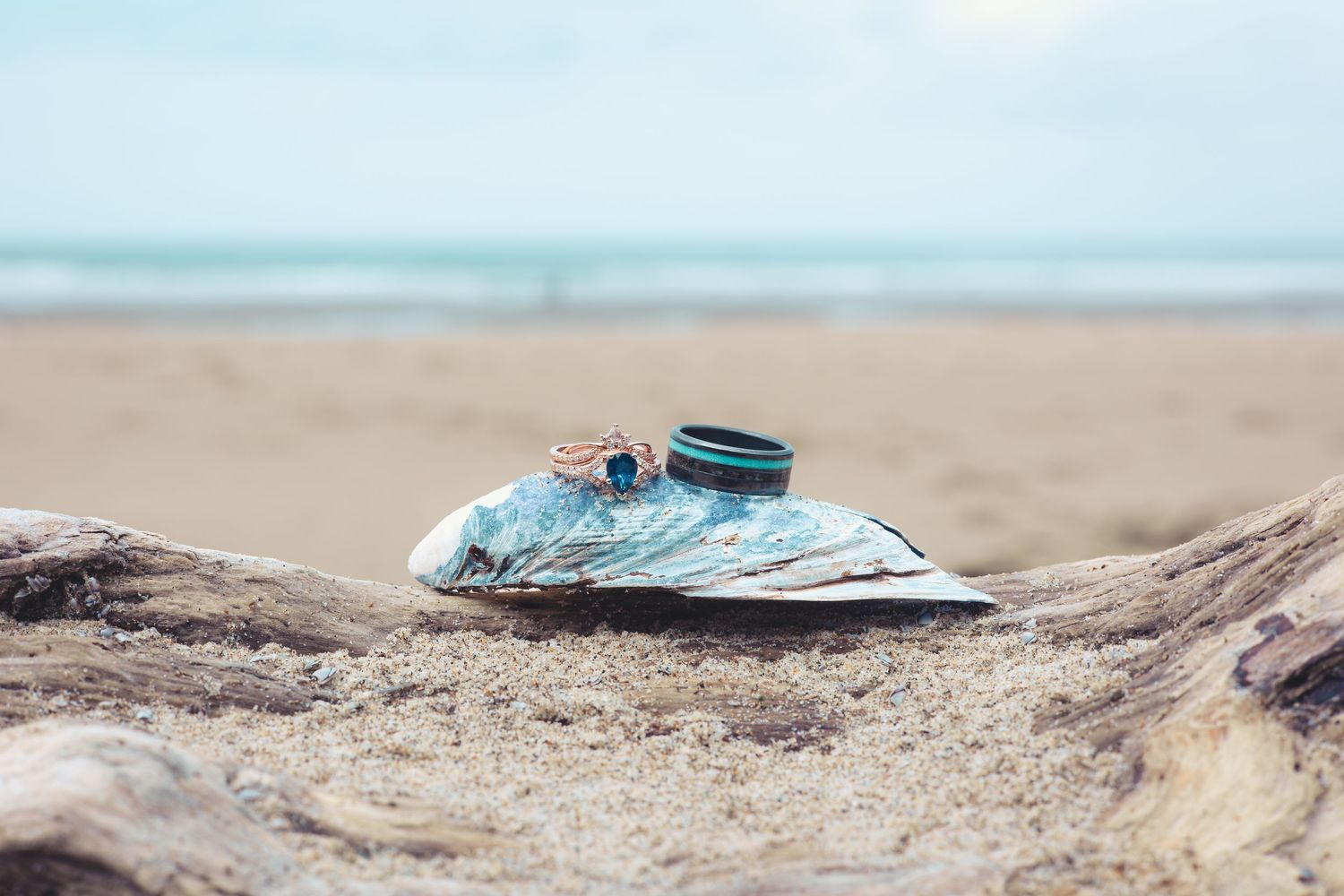 Two wedding rings resting on a blue fabric atop driftwood on a sandy beach.