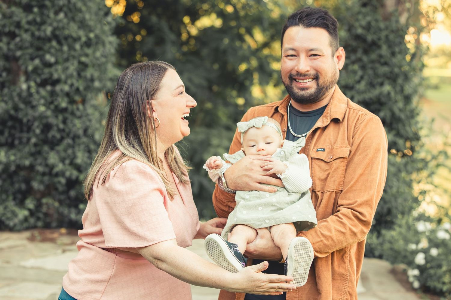 Joyful parents laughing and playing with their baby girl outdoors in a beautiful green garden setting.