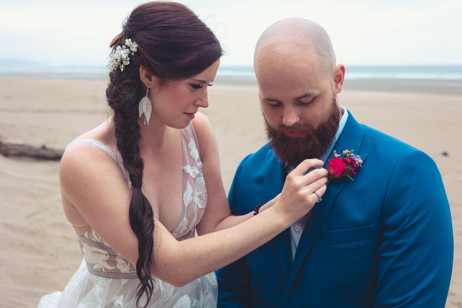 Bride with braided hair pins boutonniere on bearded groom in blue suit on beach.
