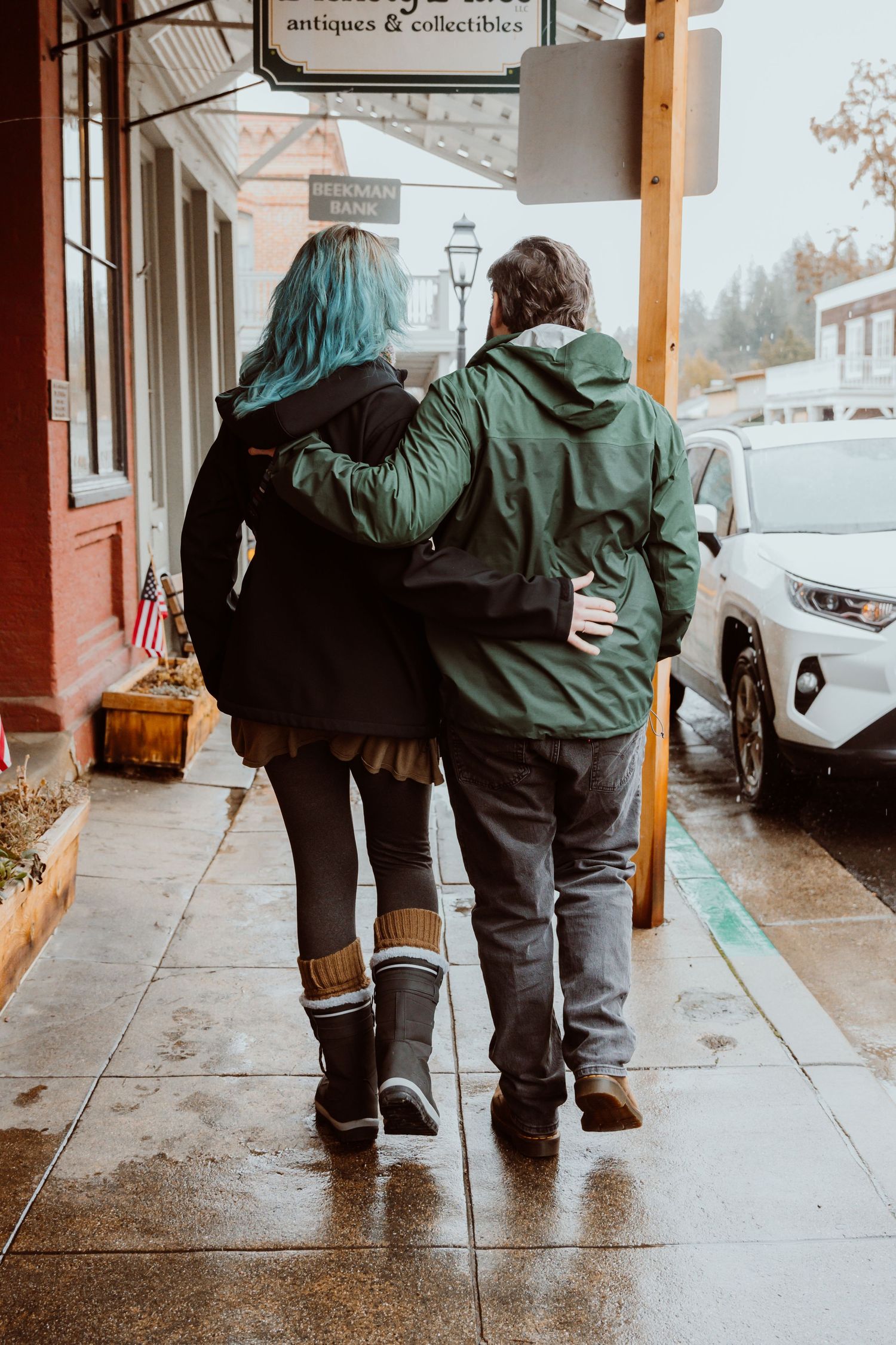 A couple walking arm-in-arm down a rainy sidewalk past antique shops.